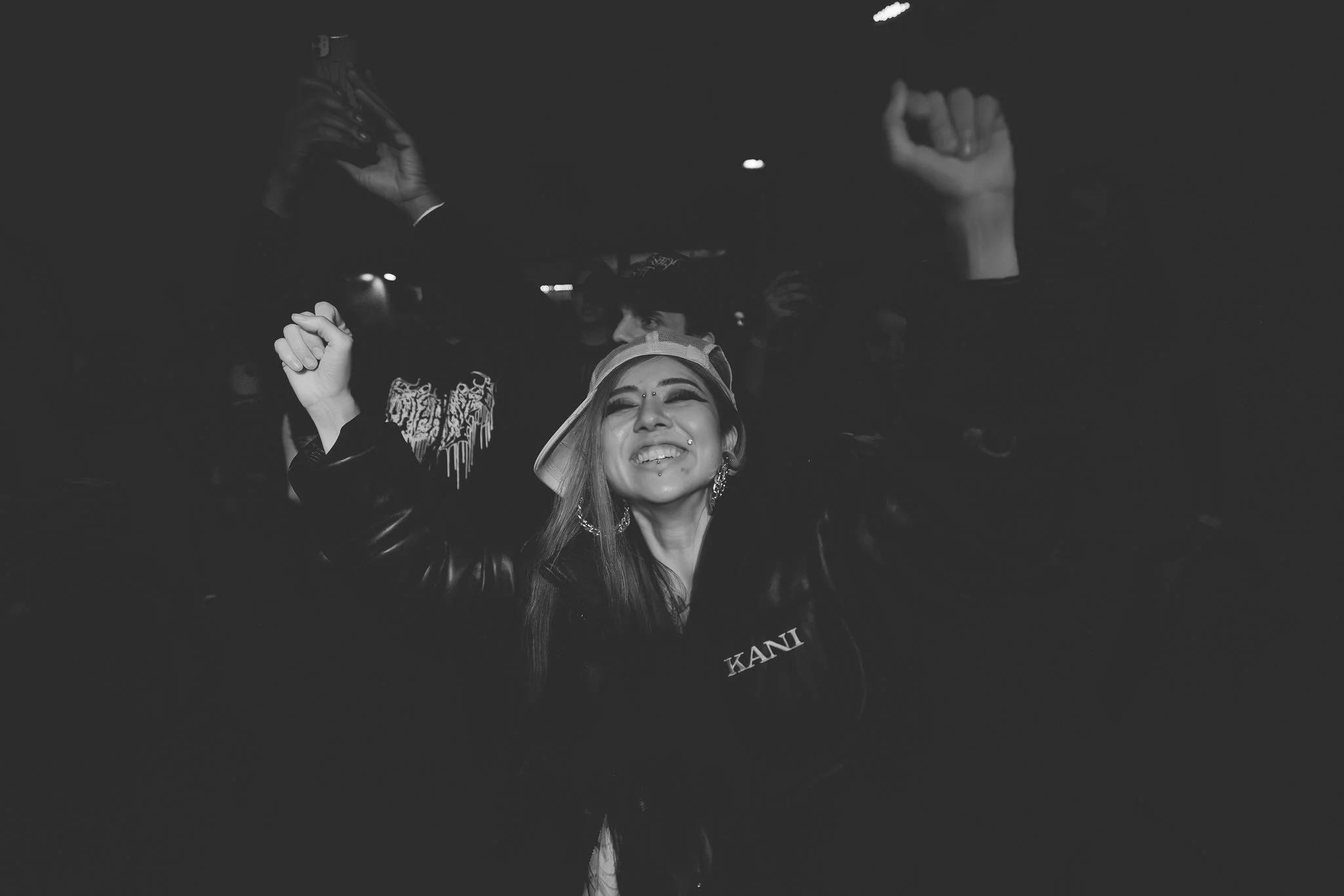 A young woman with long hair, a septum piercing, and earrings, smiling with her eyes closed, wearing a backwards cap and a jacket with the word 'KANI' on it, raising her arms in a joyful pose in a dark environment.
