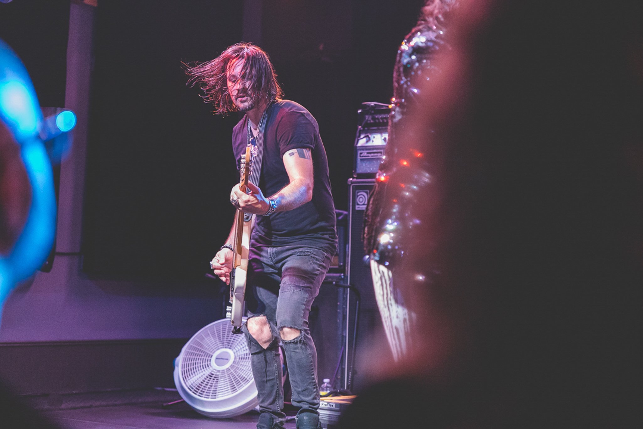 A male musician with long hair playing an electric guitar on stage, wearing a black t-shirt, ripped jeans, and several wristbands, with stage equipment and a mirror behind him.