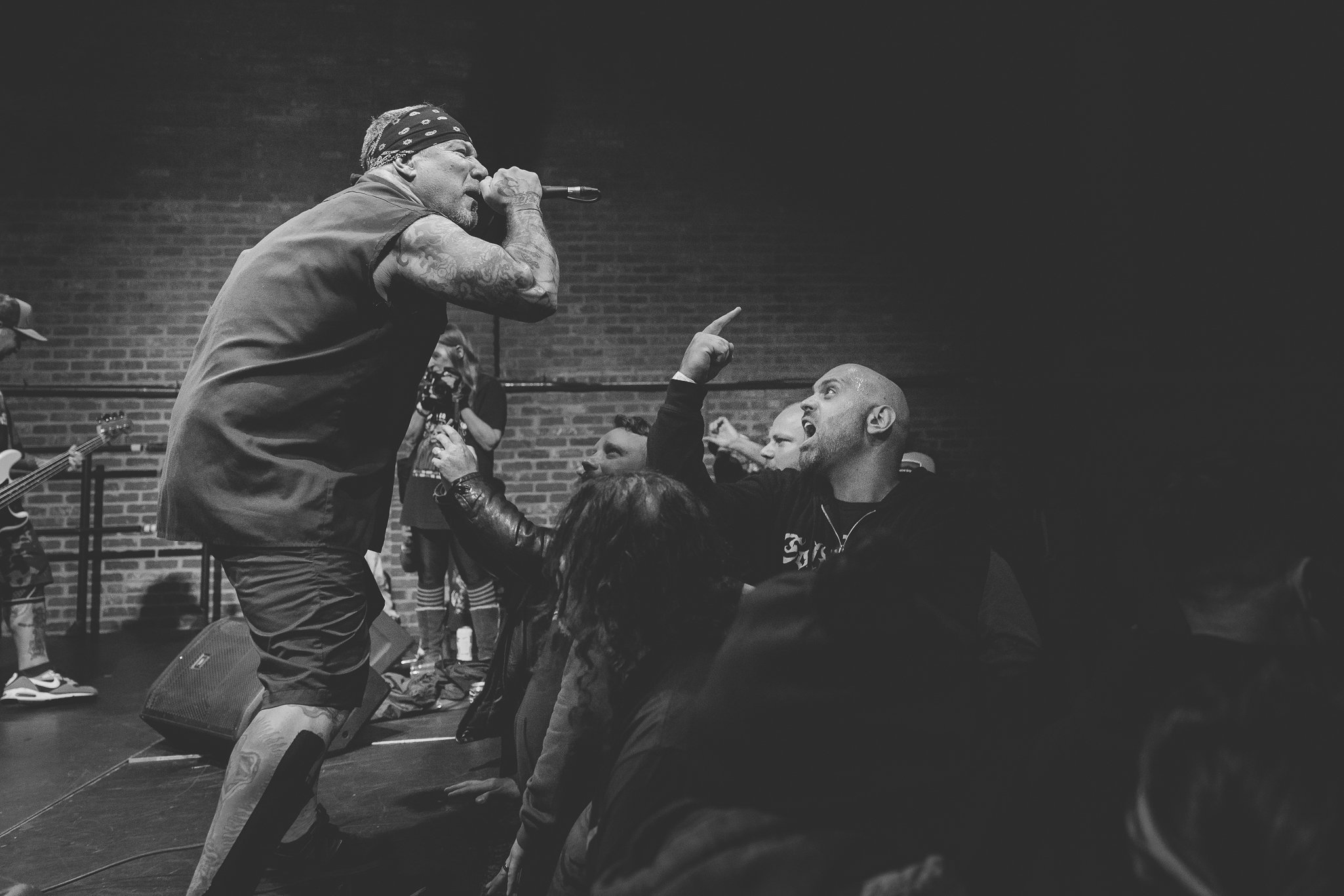 A black-and-white photo of a man wearing a bandana and sleeveless shirt singing into a microphone, interacting with an audience member who is pointing back at him in an indoor setting with a brick wall background.