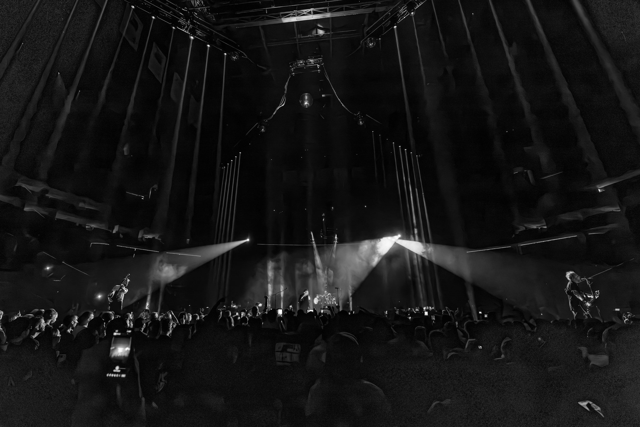 A black and white photo of a live concert. The stage has bright spotlights shining downward, and there are silhouettes of band members performing. The audience is visible in the foreground, watching the performance.