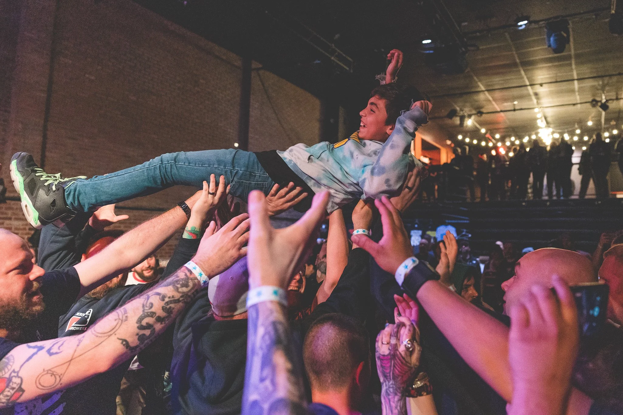 A young boy crowd surfing at a concert or music event, with people around him holding him up and stage lights overhead.