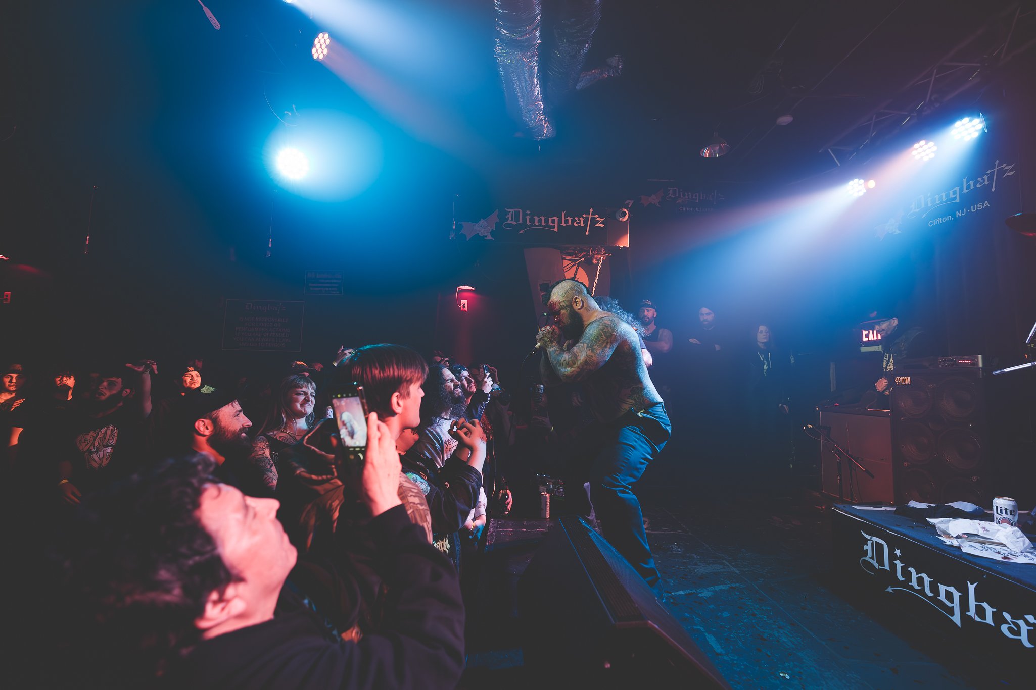 A live music concert with a shirtless, heavily tattooed performer singing into a microphone on stage. The audience is engaged, with some taking photos. The stage is illuminated with blue and purple lights, and a sign with "Dingbatz" and "Clifton, NJ 