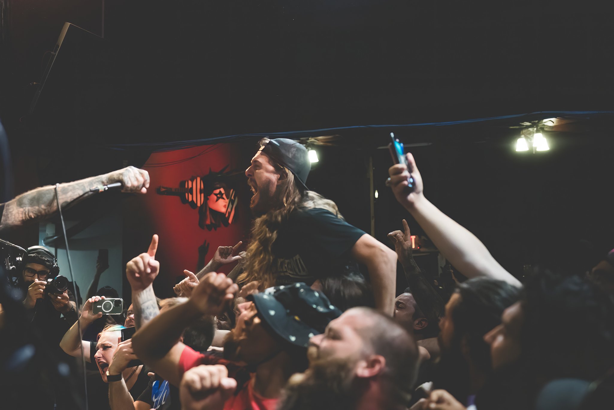 A woman with long hair and a cap on her head at a concert or event, surrounded by a crowd, reaching out and interacting, with a man recording the moment with a camera nearby, and people holding up their phones.