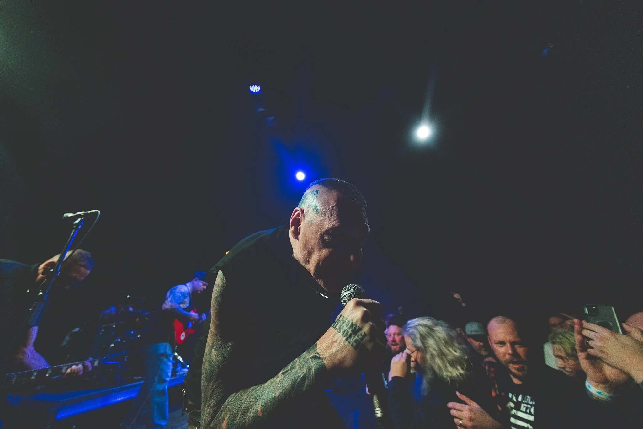 A group of people gathered at a concert or event, with a focus on a man holding a microphone, surrounded by others who appear to be singing or enjoying the performance, and a dark stage illuminated with blue and white lights overhead.