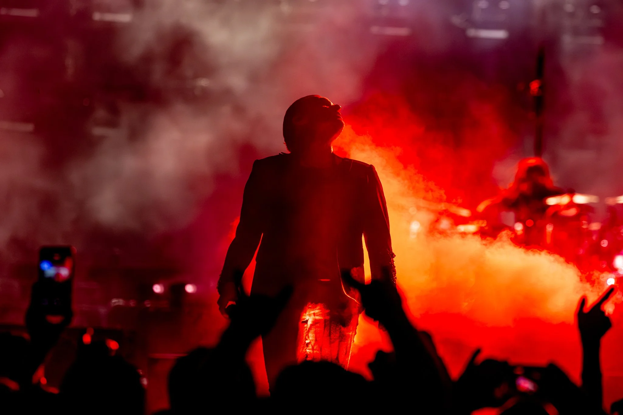 A silhouette of a person standing in front of red smoke and flames at night, with people and phones visible in the foreground.