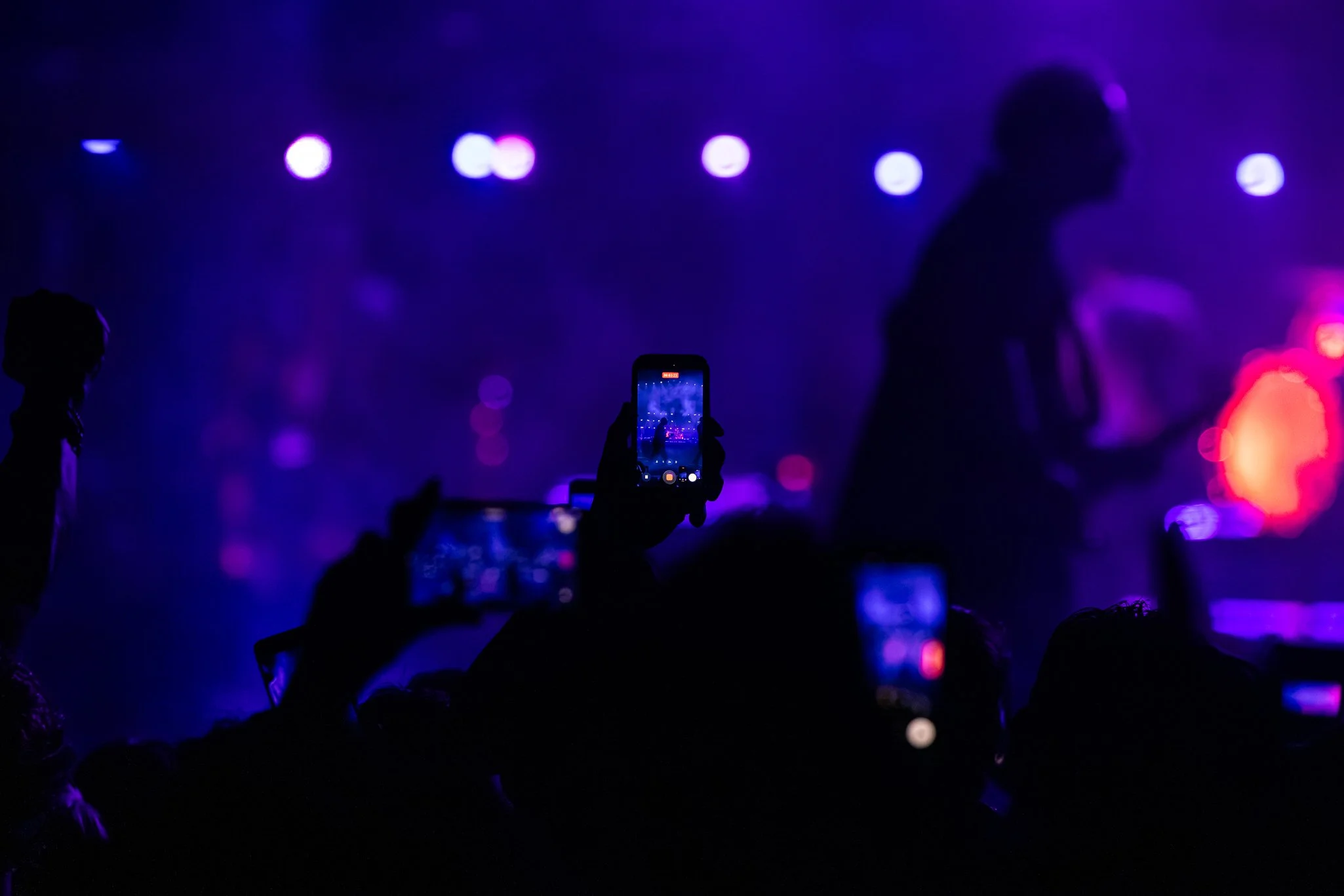Silhouettes of people at a concert or event taking photos of the stage with mobile phones, illuminated by purple and pink stage lights.
