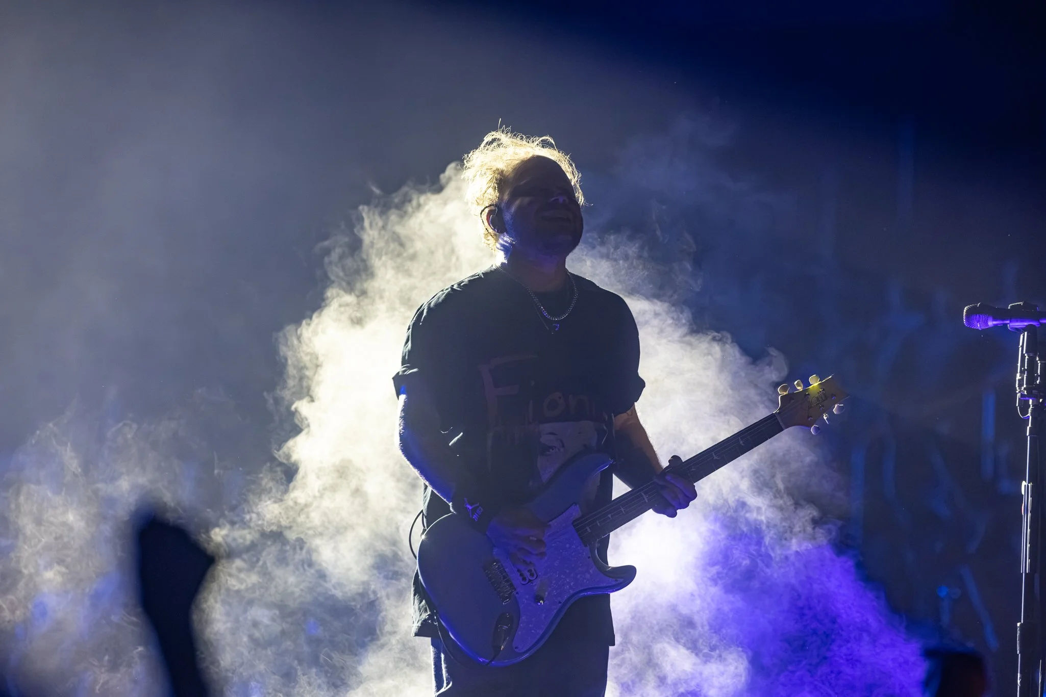 A musician playing an electric guitar on stage surrounded by smoke and blue lighting, with a microphone stand nearby.