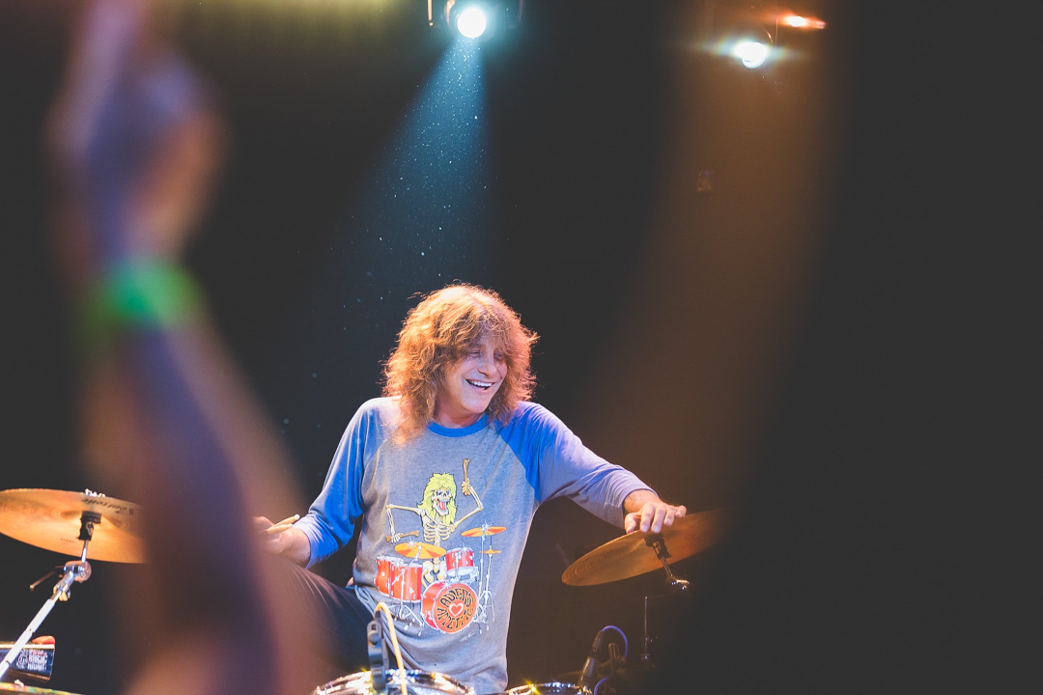 A drummer with long curly hair smiling while playing drums on stage, illuminated by colorful stage lights in the background.