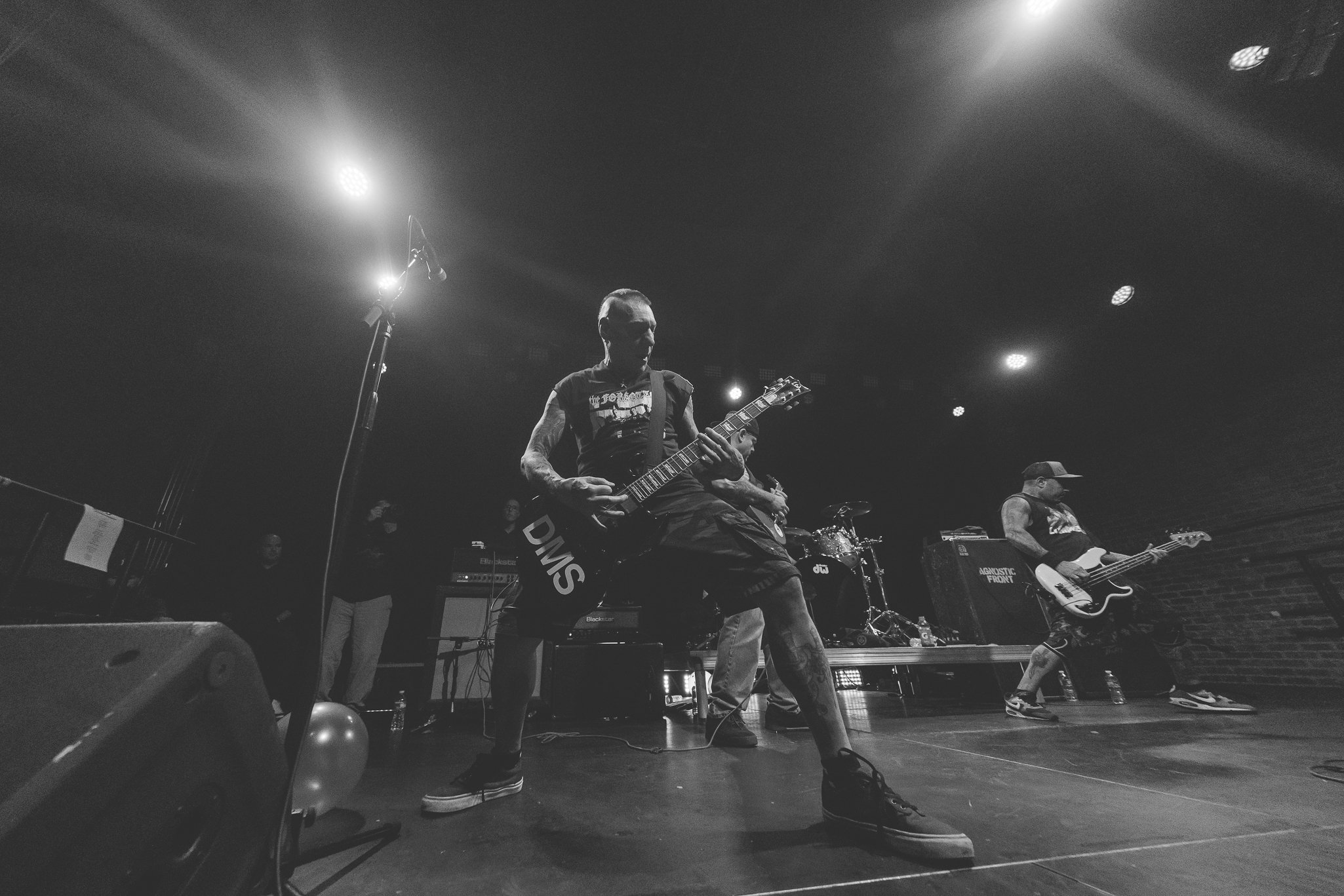 Black and white photo of a live music performance with multiple musicians on stage, including a guitarist in the foreground and others playing instruments in the background, stage lights overhead.