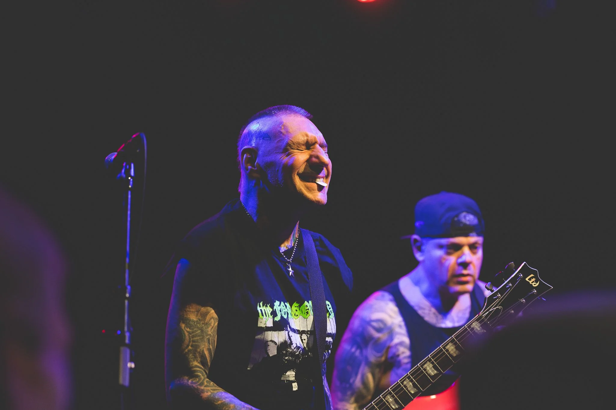 A man with a mohawk haircut and tattoos on his arms, smiling while performing with a band on stage, wearing a graphic t-shirt, and playing an electric guitar.