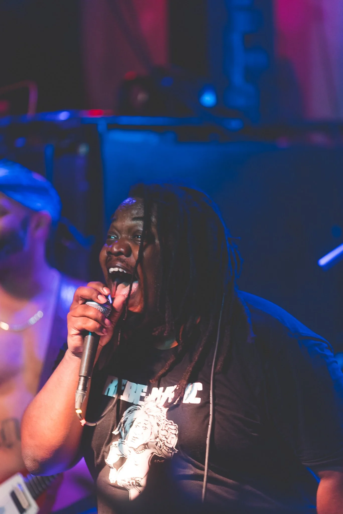 A person singing into a microphone at a nightclub with blue and purple lighting, wearing a black t-shirt with a lion graphic.