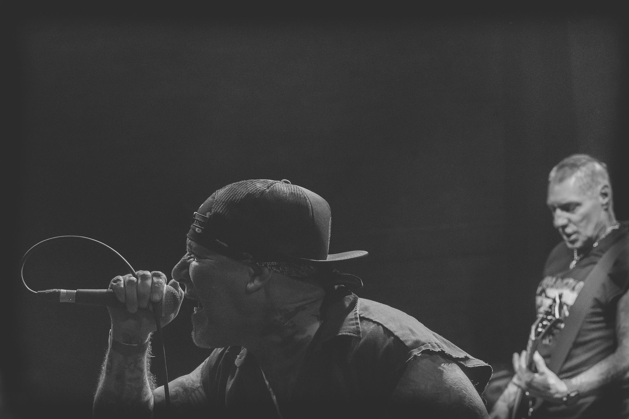 A black and white photo of two men, one singing into a microphone and the other standing nearby, both wearing casual clothing and caps, on a stage or in a performance setting.