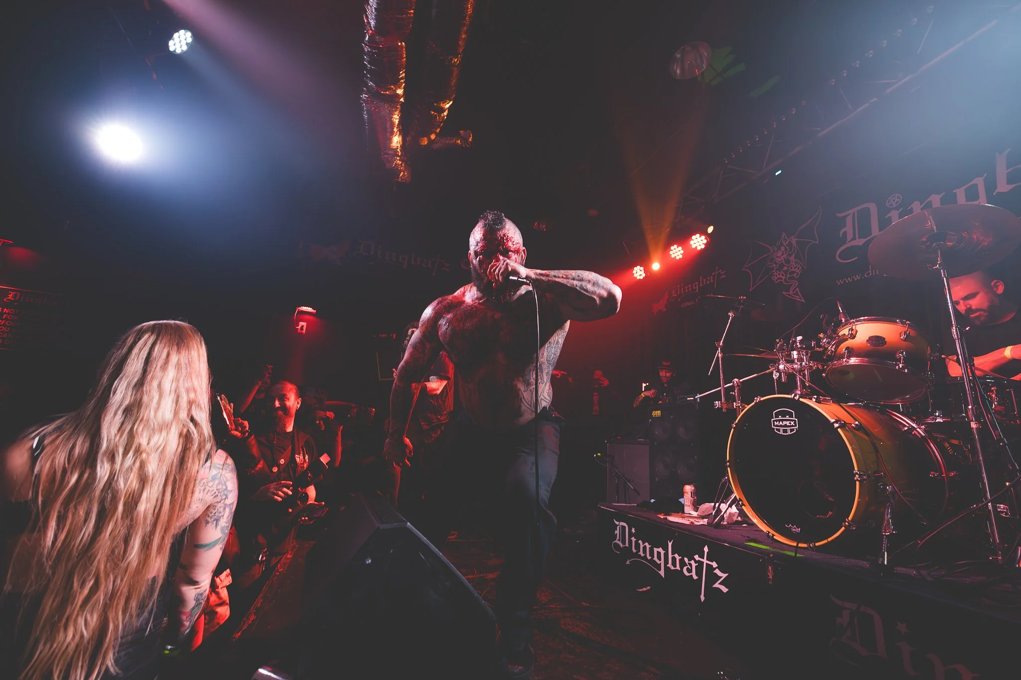 A shirtless man singing into a microphone on stage during a concert with a band and audience in front. The stage has red and white lighting, a drum set, and a backdrop with the band's logo.