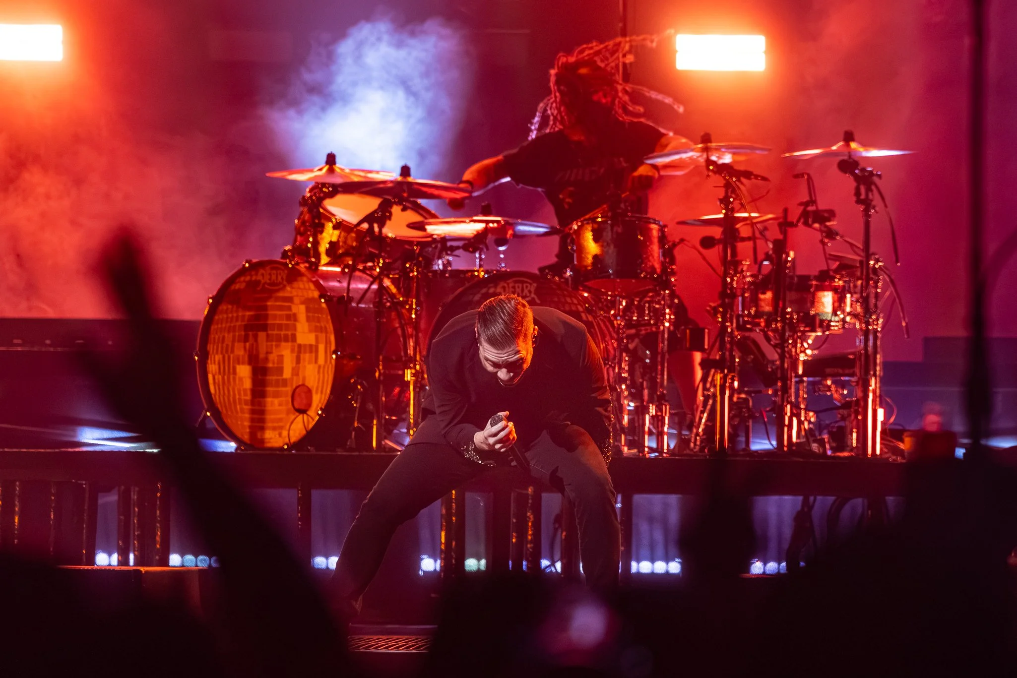 A performer dressed in black is crouching with a microphone in hand, singing passionately on stage. Behind them, a drummer with dreadlocks is playing a drum set illuminated by red and orange stage lights, with smoke and bright lights creating a dynam