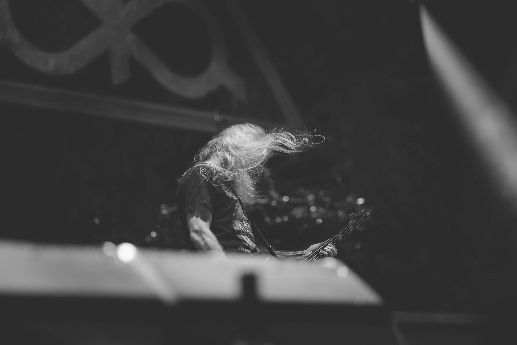 A person with long, curly hair playing an electric guitar on stage at night, with bright stage lights and blurry audience lights in the background.