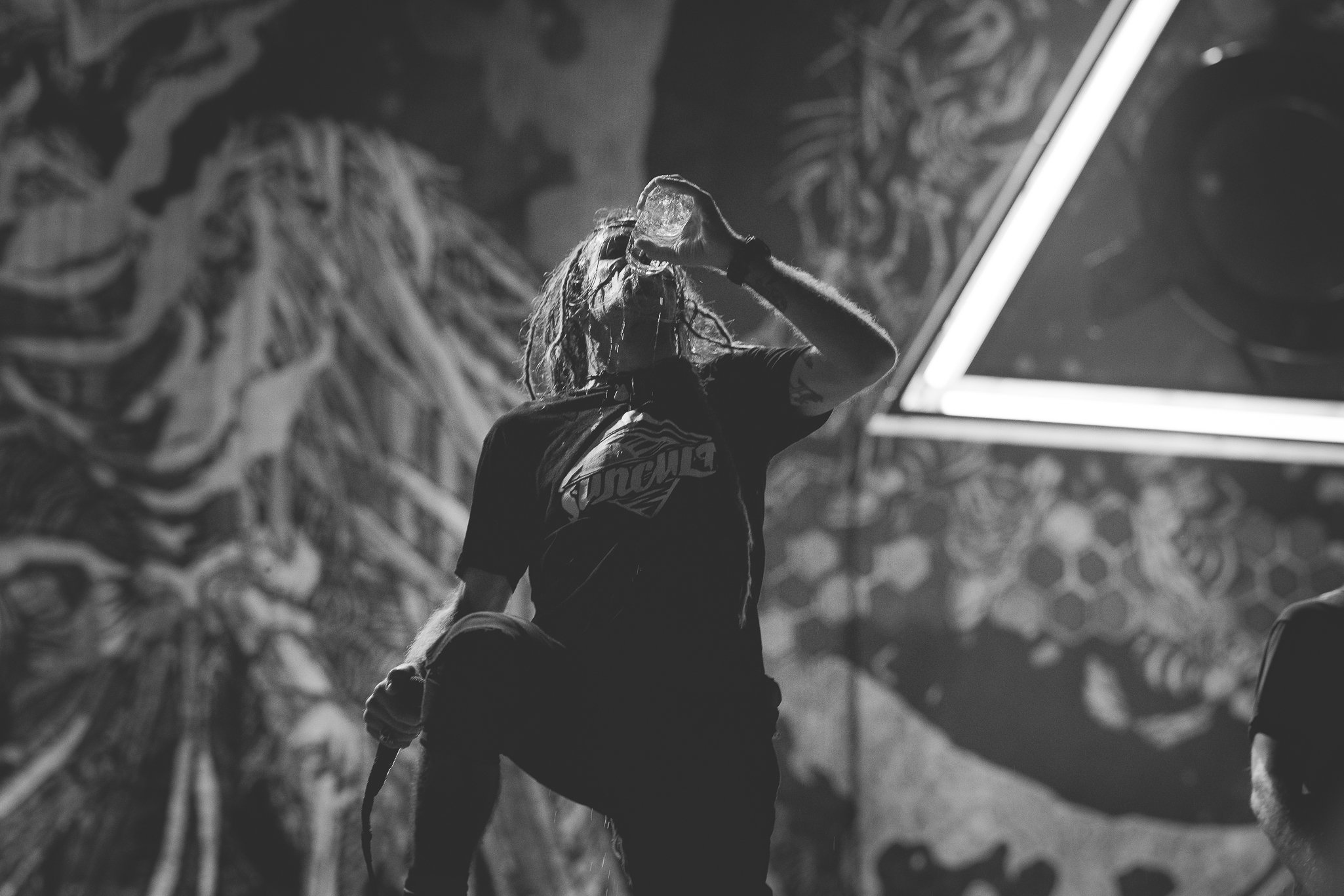 A person with long hair, wearing a Black T-shirt, is holding a water bottle and drinking water. The person appears to be in an indoor climbing gym with graffiti-style artwork on the wall in the background.