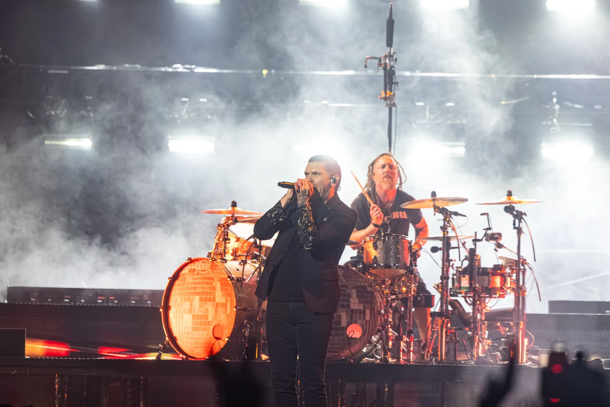 A singer performs on stage with a microphone, wearing a black outfit, with a drummer in the background. The stage is filled with smoke and dramatic lighting.