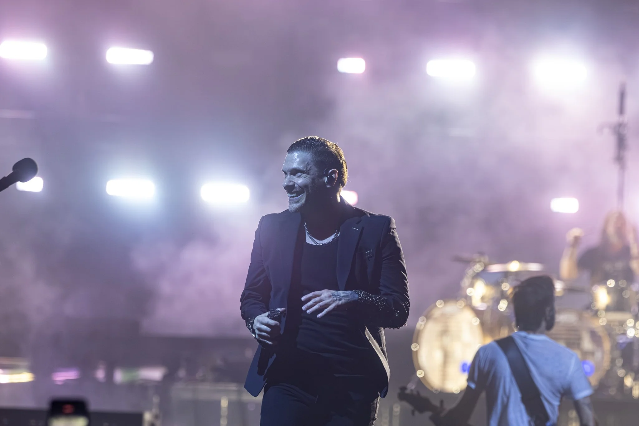 A man in a black suit and jewelry smiling with his hand on his stomach on stage with bright lights and a band in the background.