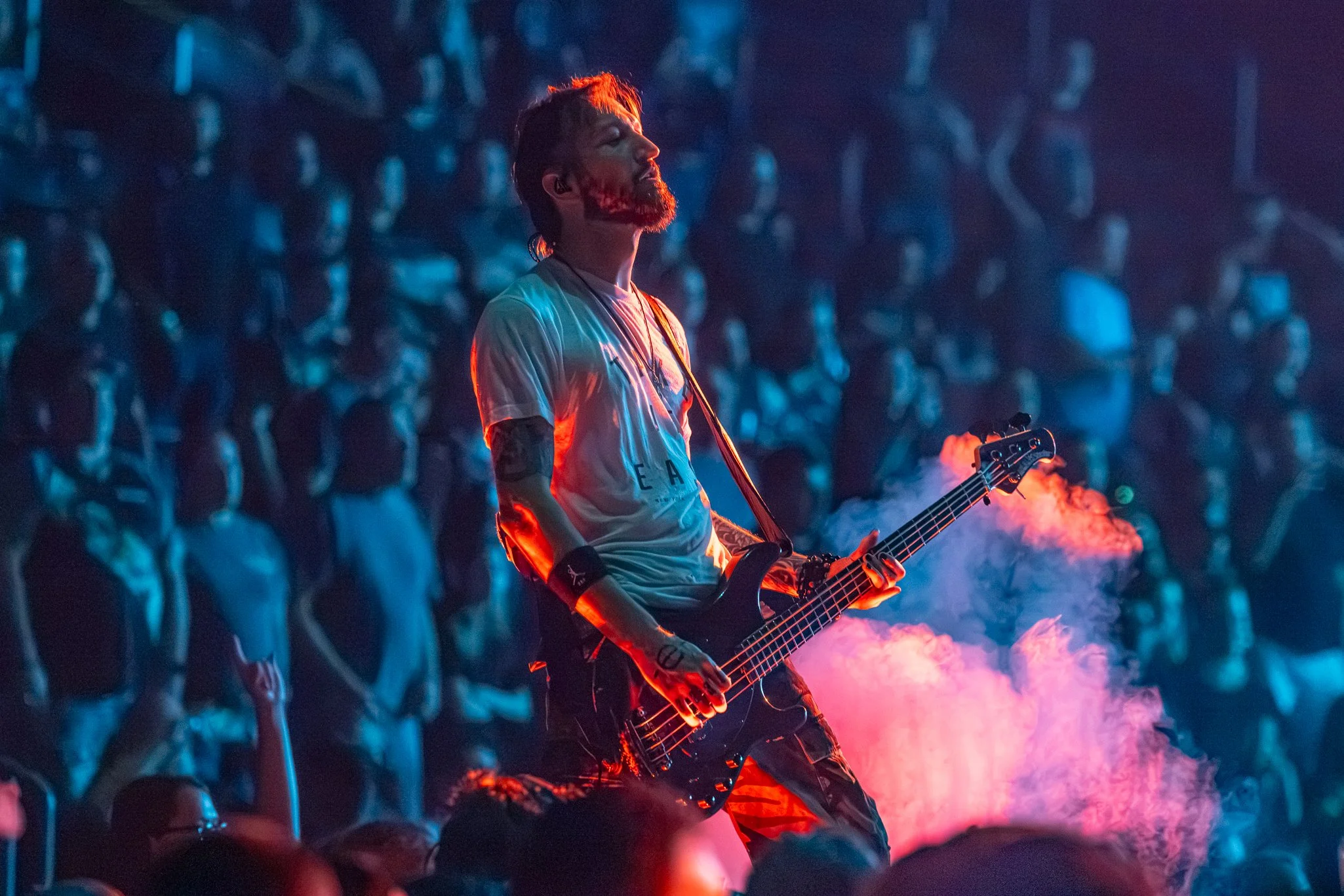 A man with a beard and styled hair playing an electric bass guitar at a concert, illuminated by colorful stage lights with an audience in the background.