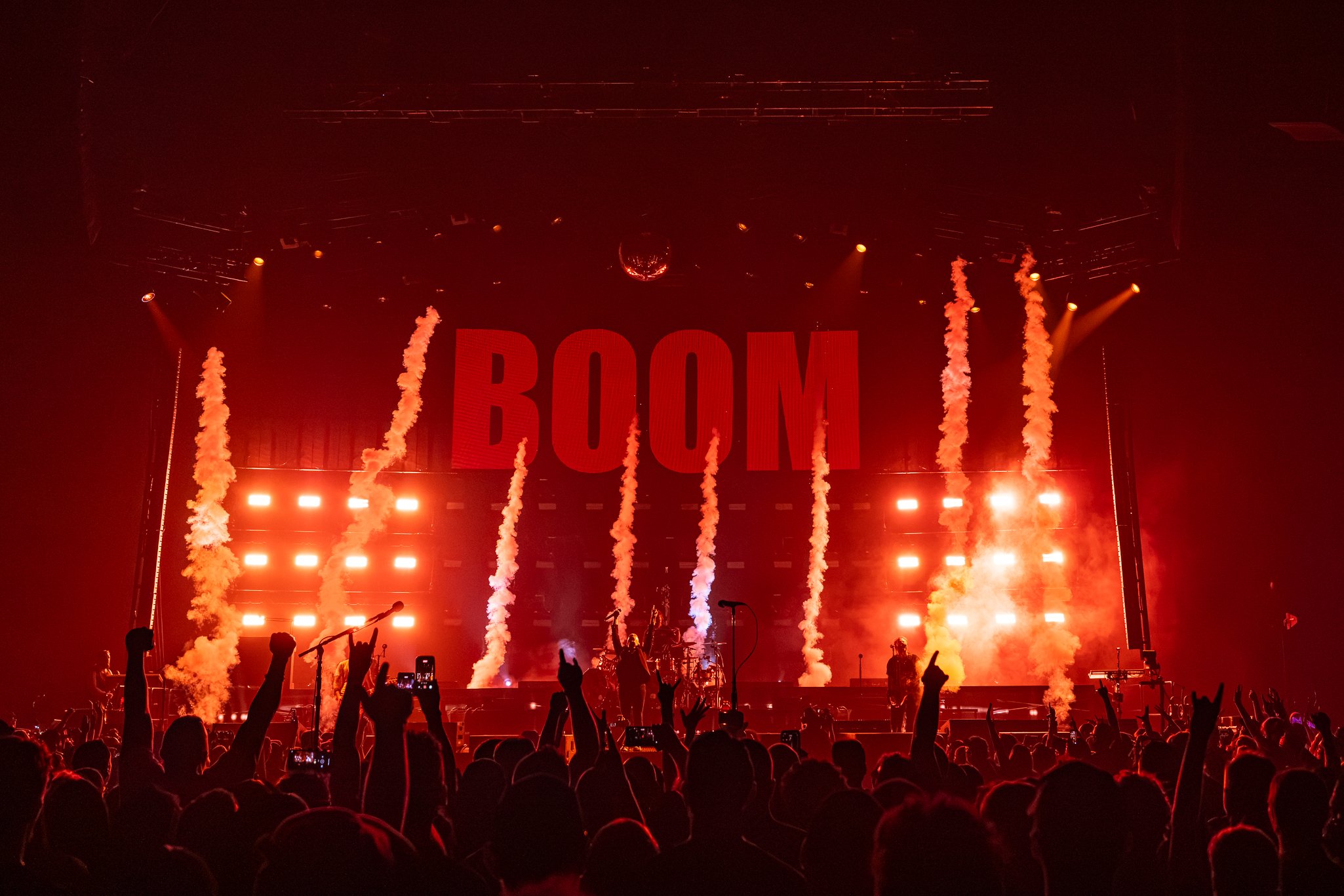 Concert scene with a large crowd, stage with bright red lights, smoke effects, and the word 'BOOM' displayed on a large screen.