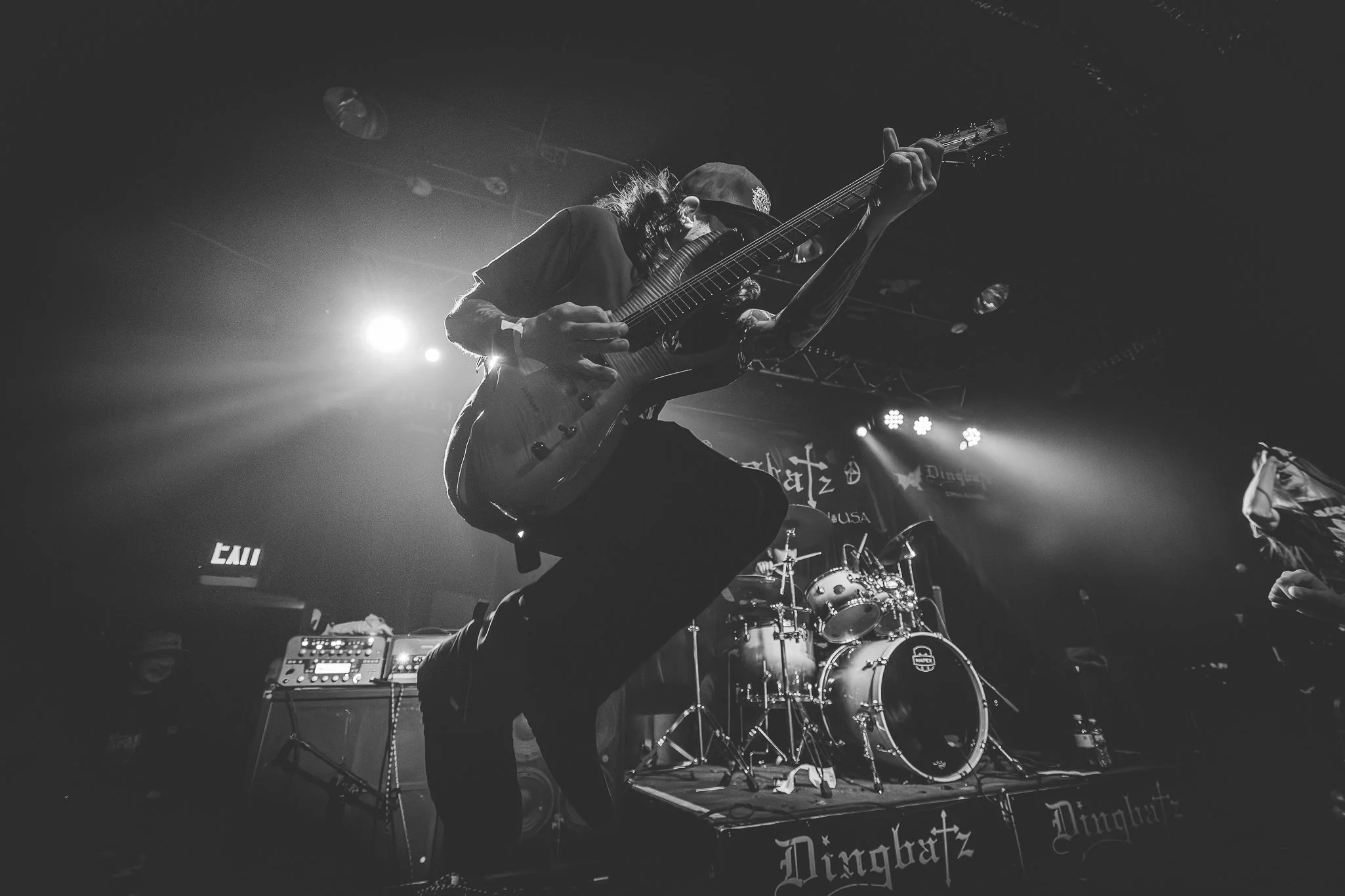 A black and white photo of a musician playing an electric guitar on stage, with stage lights and a drum set in the background.