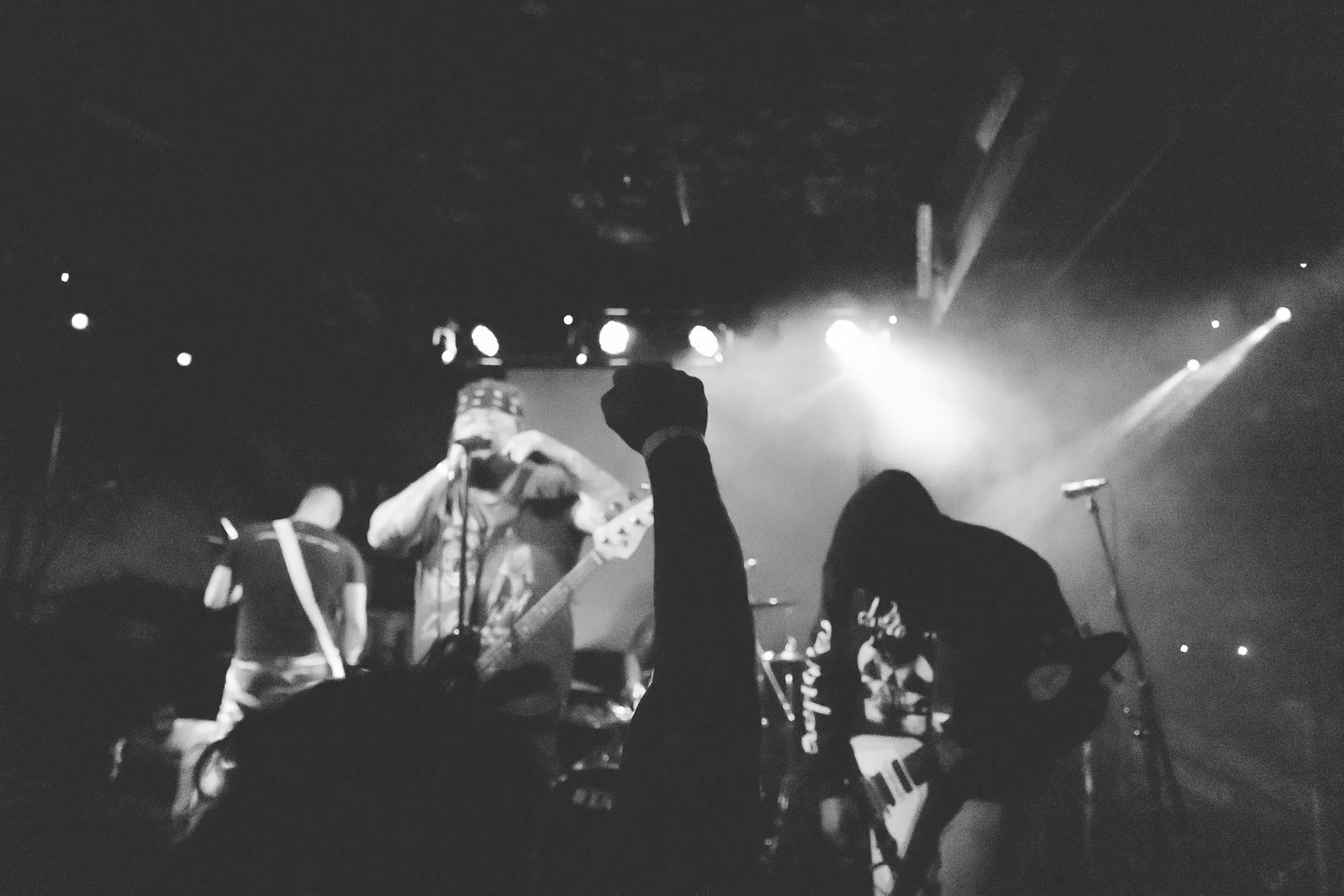 Black and white photo of a concert scene with a musician playing a guitar on stage, audience members visible with their hands raised and one person taking a photo. Bright stage lights are shining down, creating a lively atmosphere.