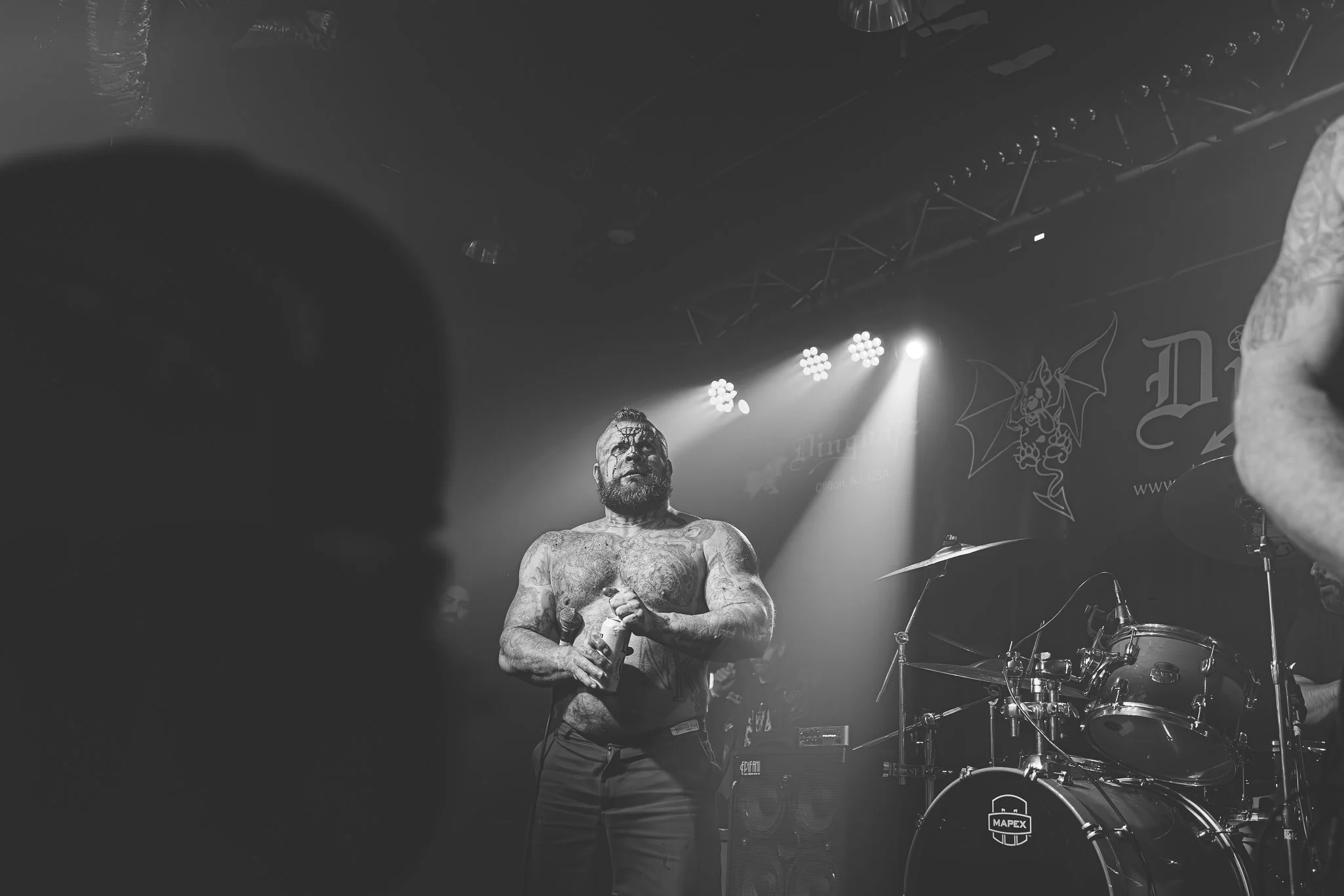 A shirtless male musician with tattoos, a beard, and face paint holding a microphone on stage during a concert, with drums and stage lighting visible in the background.