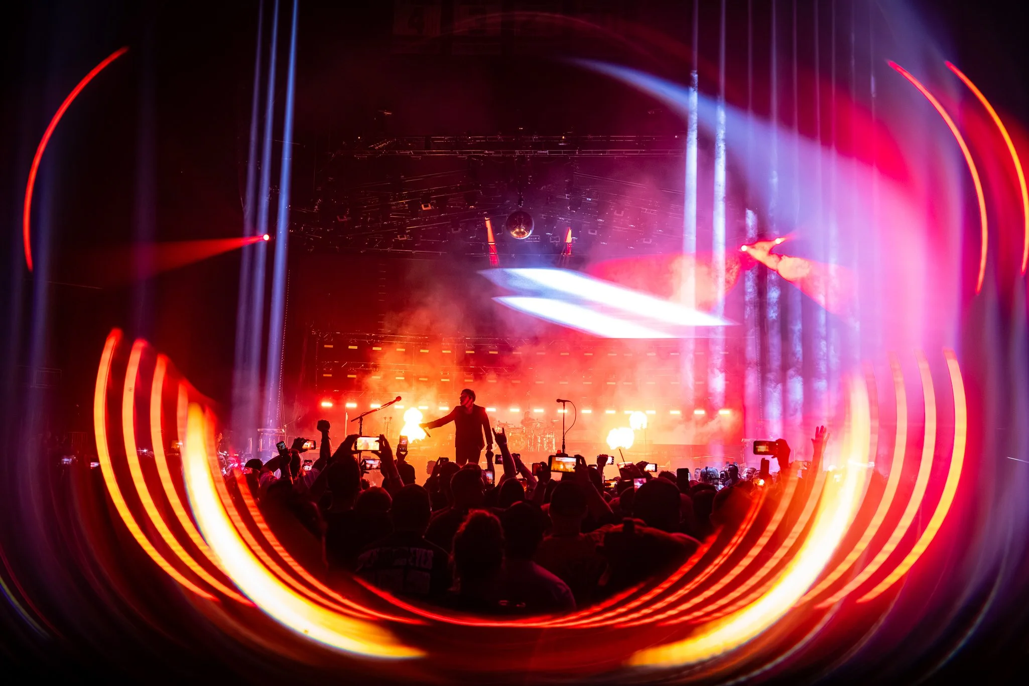 A concert stage with colorful lighting effects, a musician performing in front of a large audience holding up their phones, and a disco ball hanging from the ceiling.