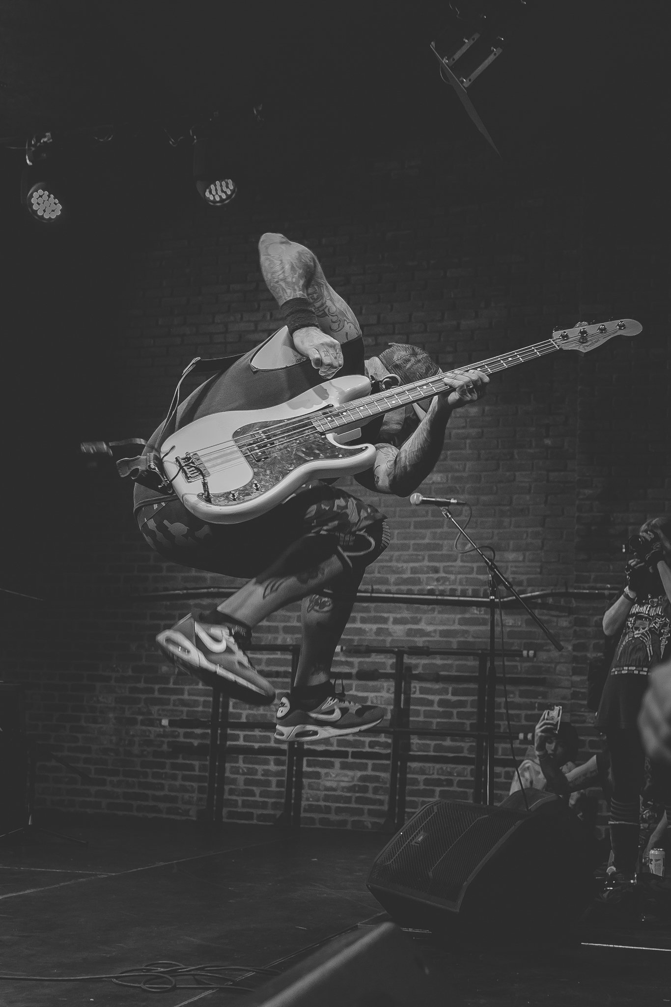 A person with tattoos jumping in the air while playing an electric bass guitar on stage in a dark venue with stage lights and a brick wall background.
