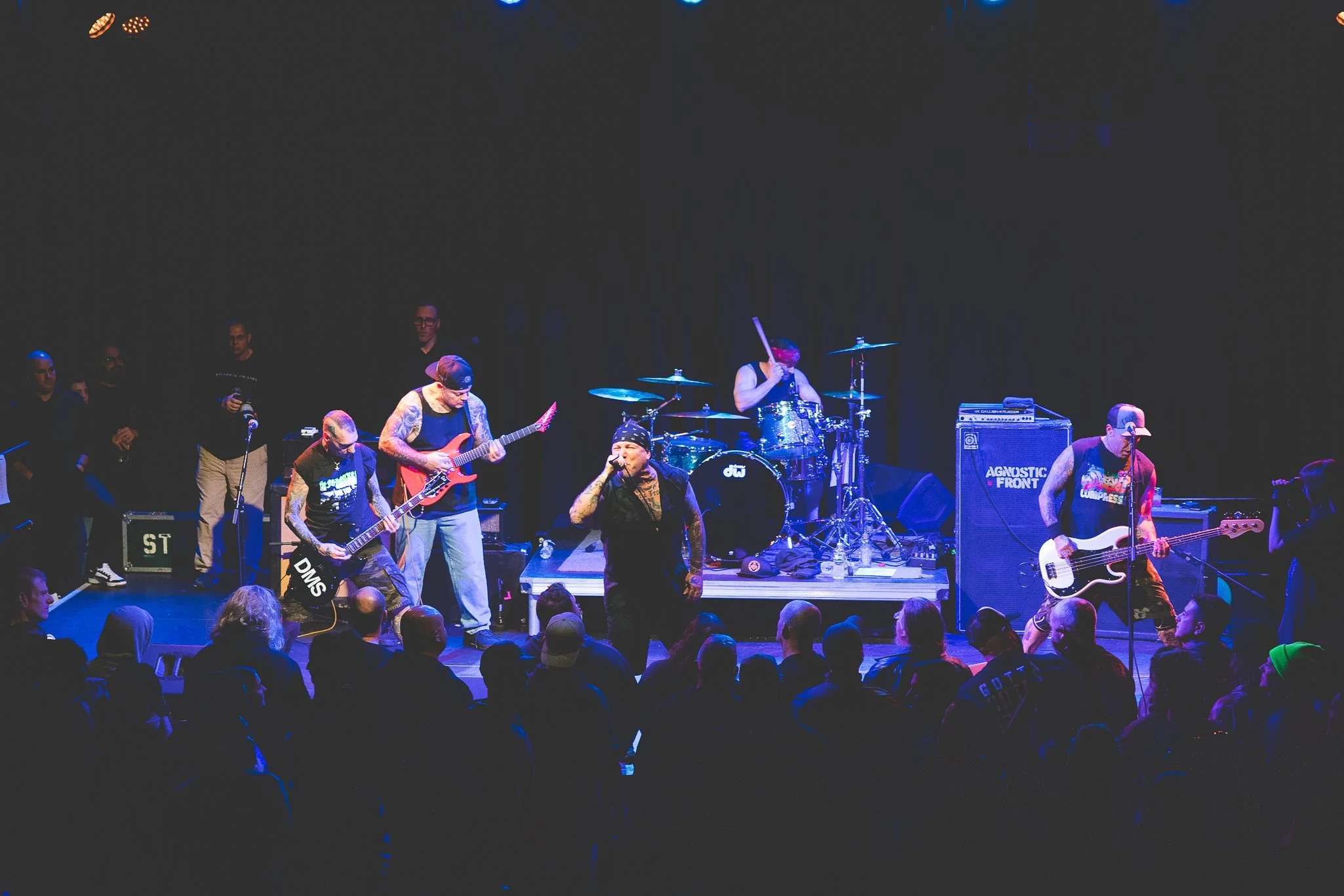 A live band performs on stage at a concert. The group includes guitarists, a drummer, and vocalists. The audience is watching from in front of the stage in a dark venue with blue lighting.