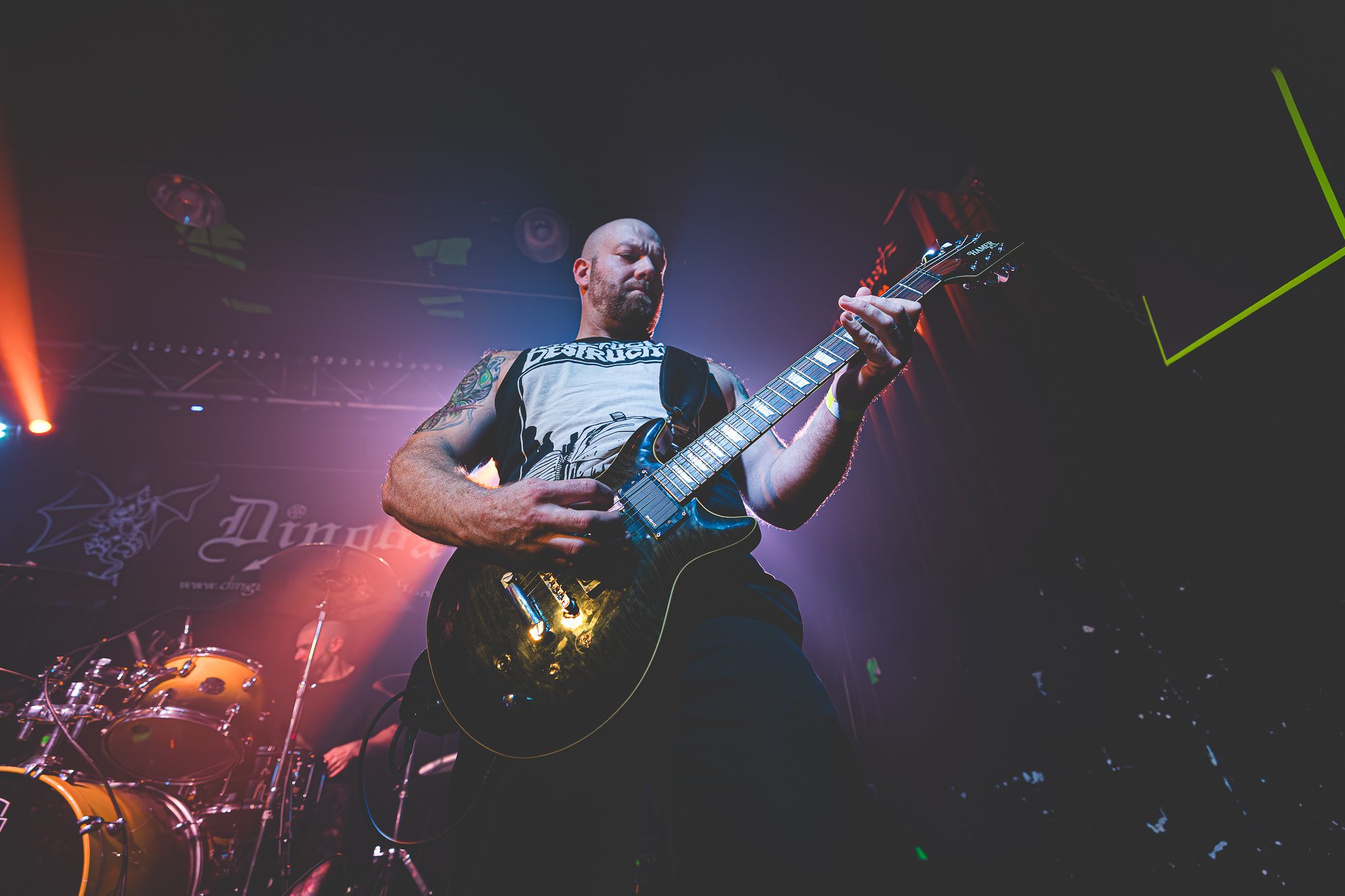 A bald male guitarist playing an electric guitar on stage with colorful lighting and a drum set in the background.