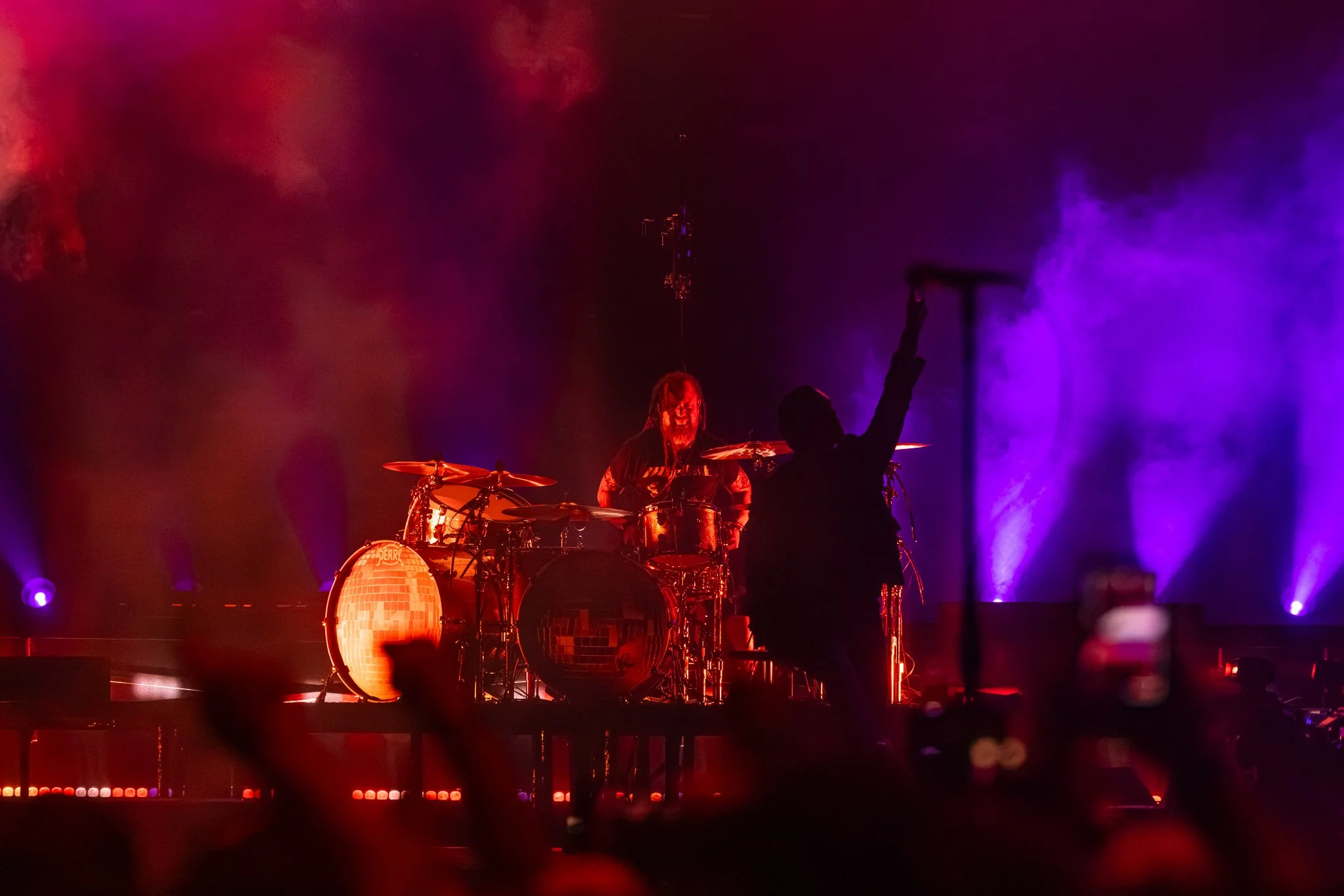 A live music performance with a drummer on stage, colorful purple and red lighting, and an audience in the foreground.