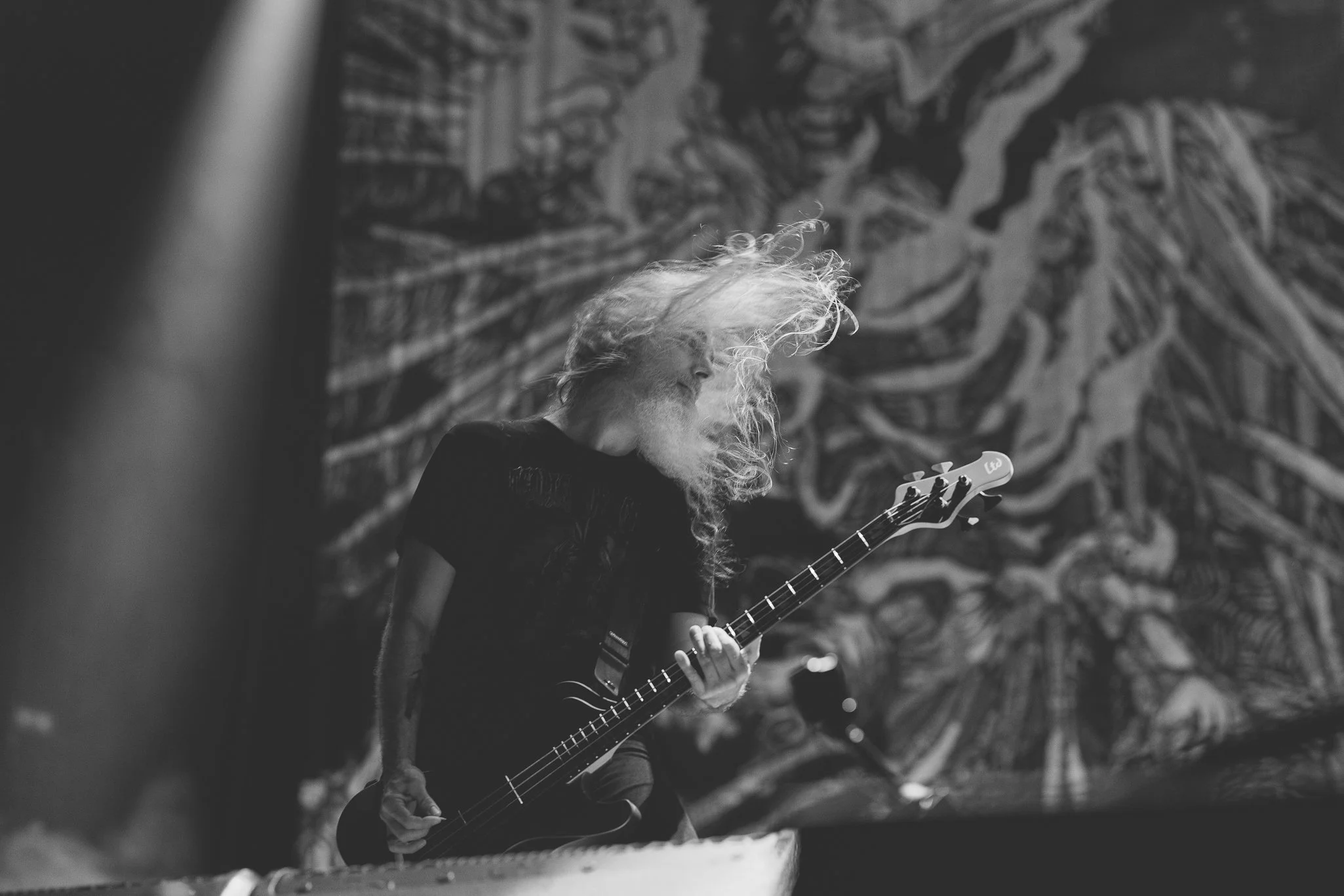 A black and white photo of a man with long, curly hair and a beard playing an electric guitar. He is facing left with a large artistic mural in the background.