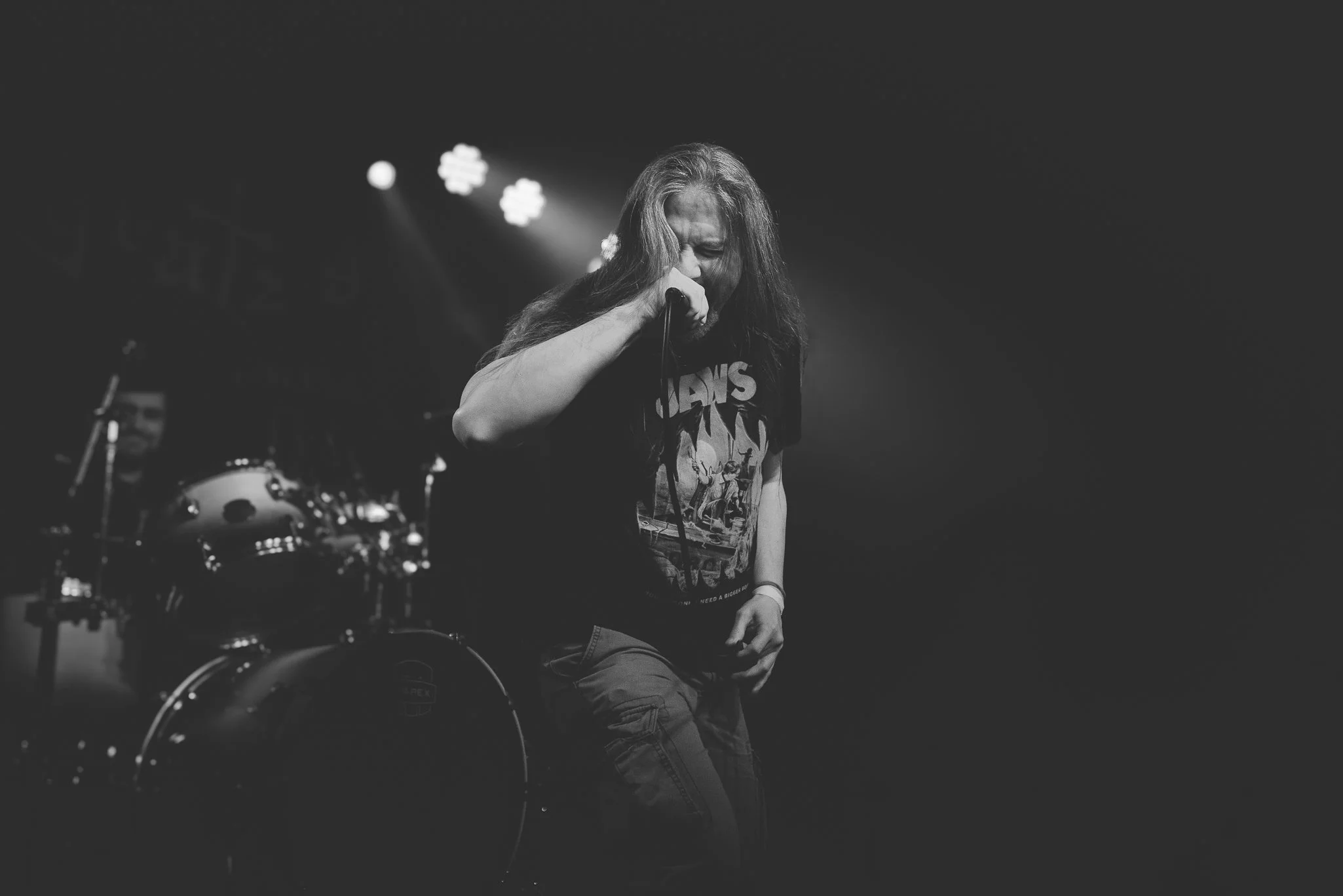 A black and white photo of a male singer with long hair, performing passionately on stage, holding a microphone close to his face. He's wearing a graphic T-shirt and jeans, with stage lights illuminating him from behind.