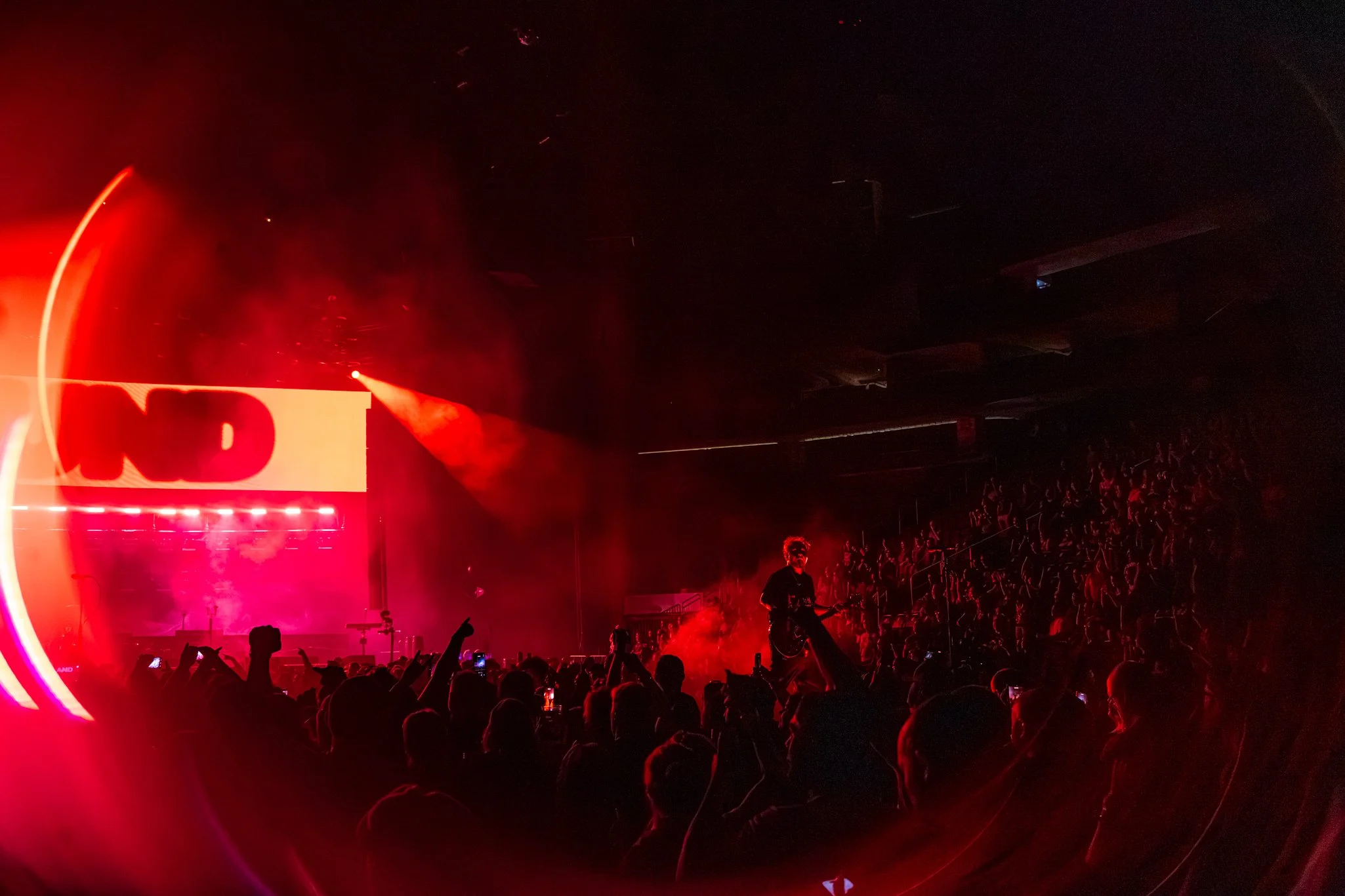 A concert scene bathed in red lighting with an audience and performers on stage. The audience is visible with some people raising their hands and taking photos.