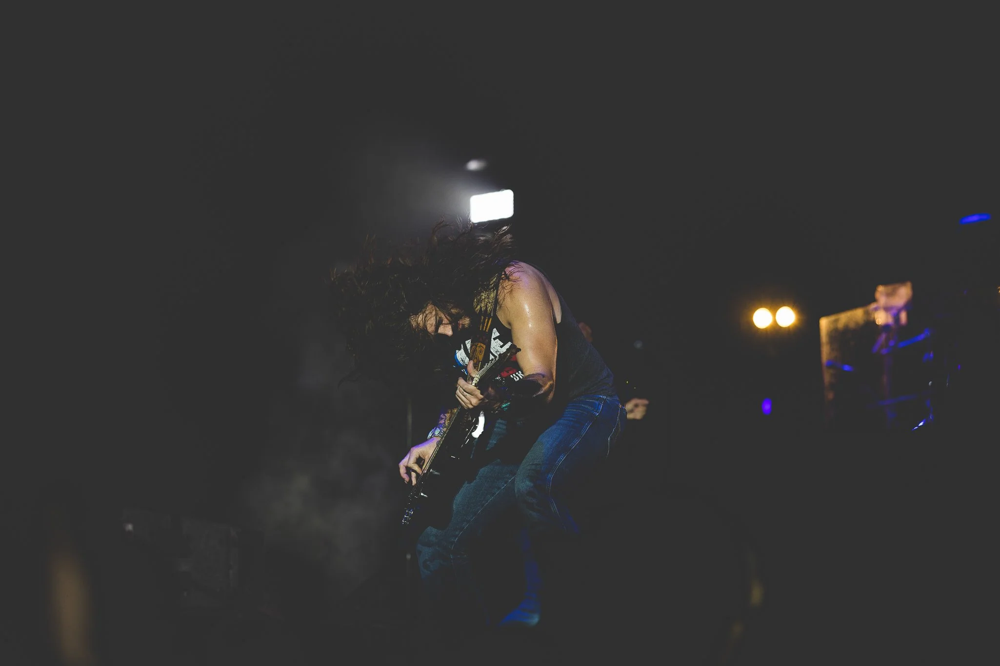 A musician with long hair playing an electric guitar on stage at night, illuminated by stage lights.