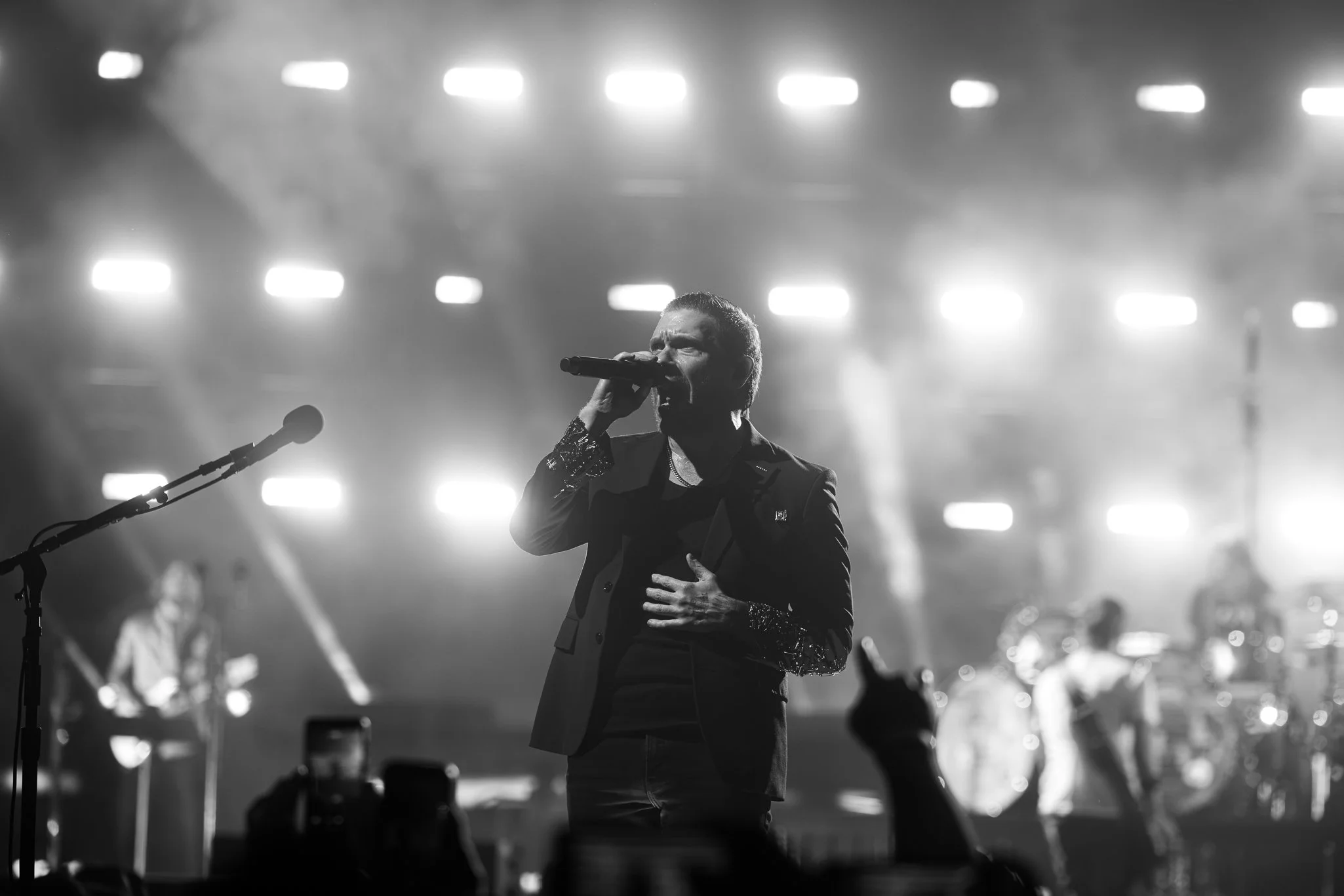 A male singer performs passionately on stage, holding a microphone with his right hand, while his left hand is placed on his chest. He is dressed in a dark blazer with sparkly sleeves and a shirt underneath. Bright stage lights and a blurred band are