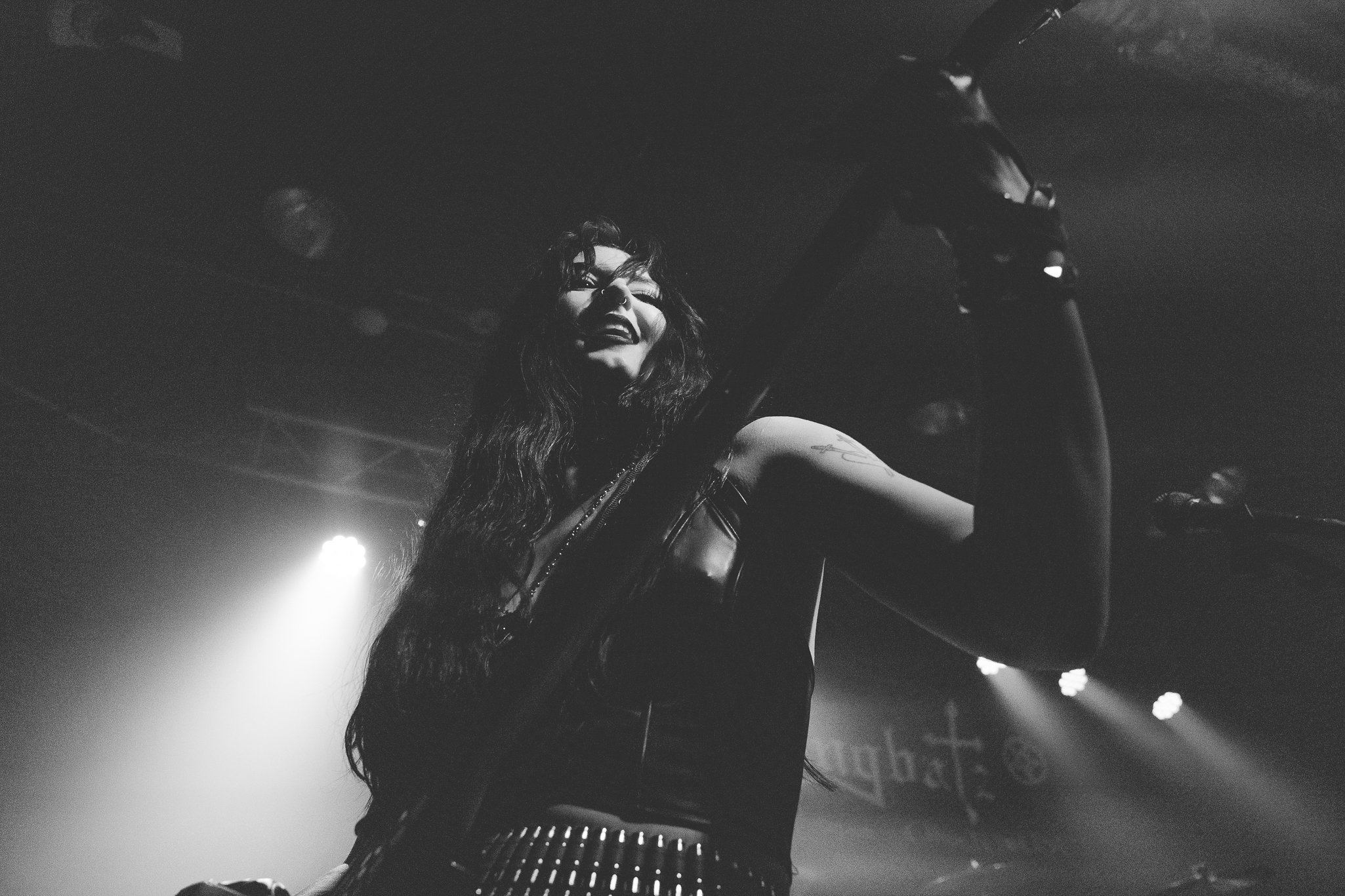 A woman with long hair and a tattoo on her arm singing and playing an instrument on stage, illuminated by stage lights.