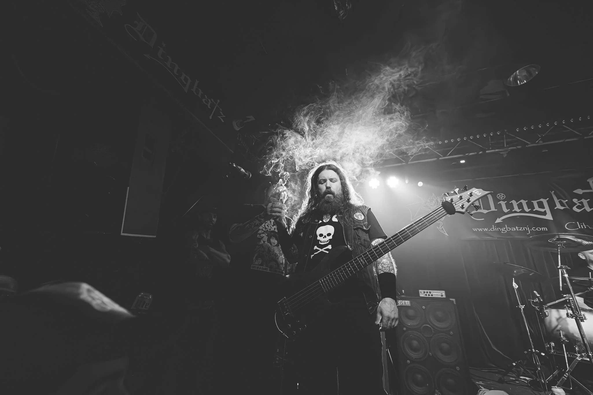 A live music performance featuring a male guitarist with long hair and a beard, wearing a skull T-shirt and denim vest, playing an electric guitar on stage. Smoke or vapor is coming from his mouth or a device, and stage lights illuminate the scene. T