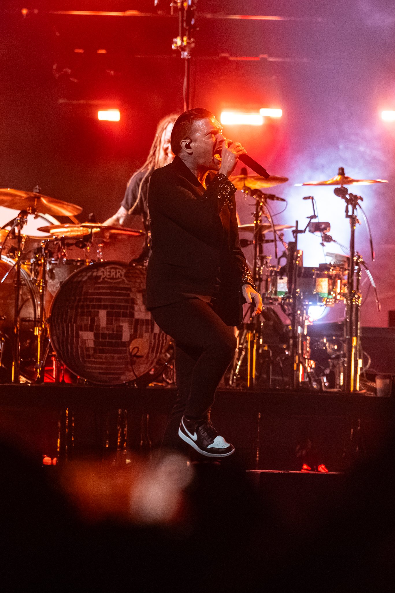 A male singer with slicked-back hair, wearing a black suit and black and white Nike sneakers, performs on stage with a microphone, illuminated by vibrant red and purple stage lights, accompanied by a drummer and musical equipment in the background.
