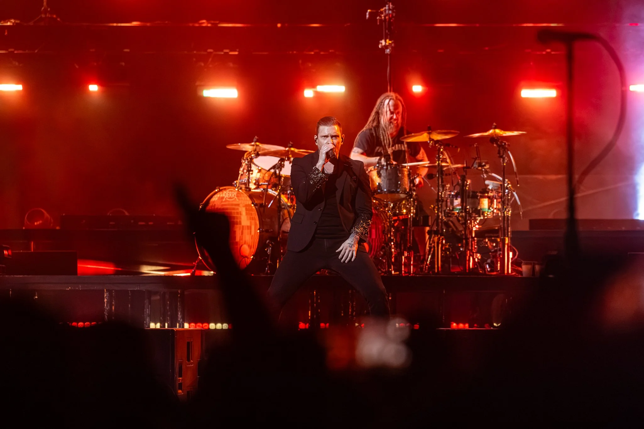 A stage with a singer in a black suit singing into a microphone, and a drummer with dreadlocks playing drums, both under red stage lights.