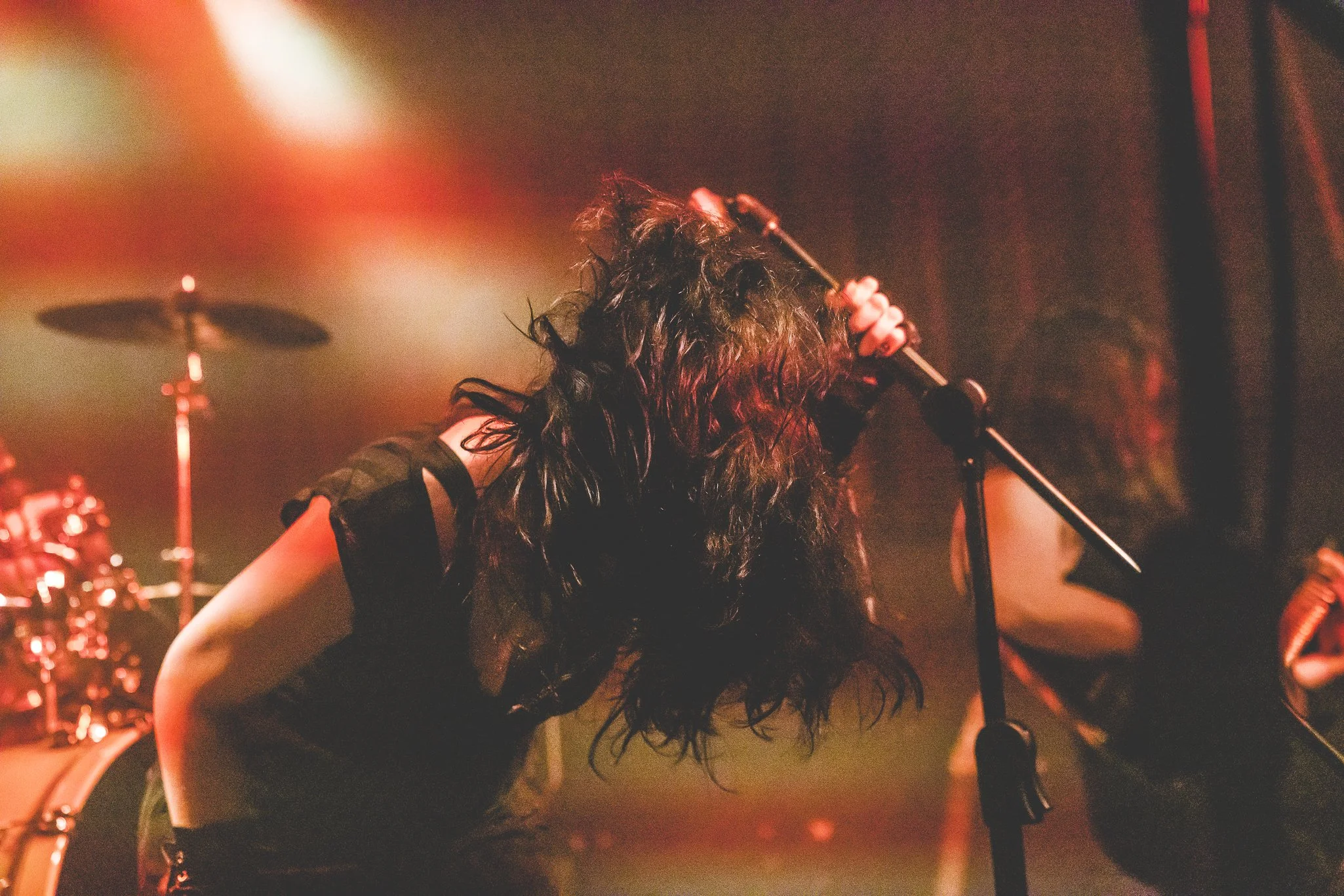 A singer with long, curly dark hair holding a microphone on stage during a concert, with warm orange and red stage lighting.
