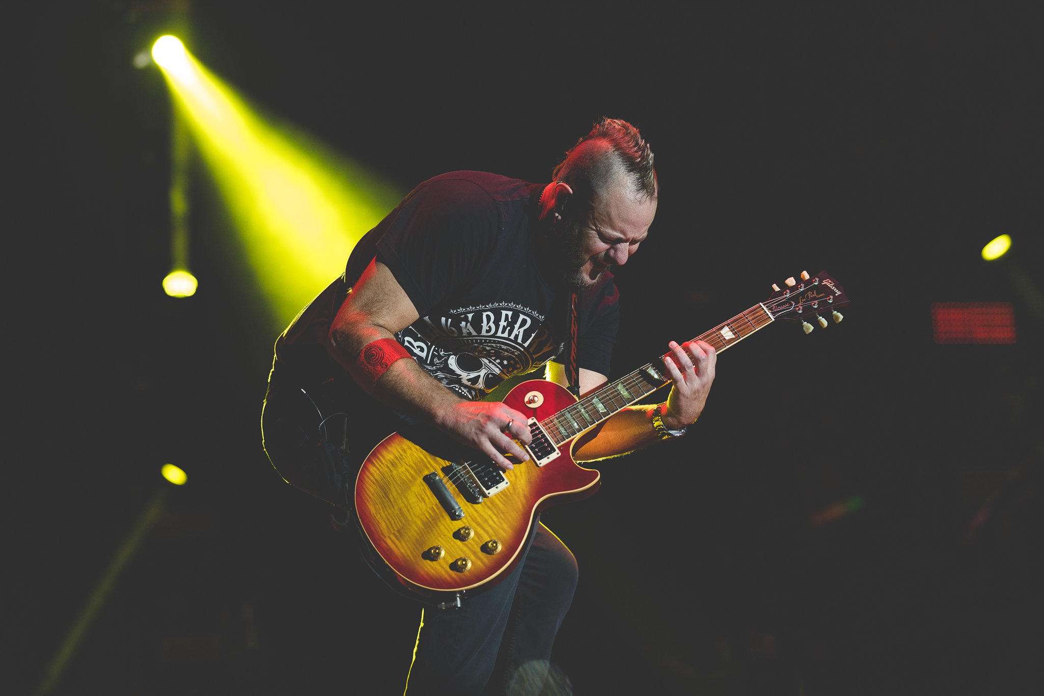 A male guitarist wearing a black t-shirt with a skull and the words 'Buckberry' and playing an orange and yellow electric guitar on stage under yellow lights.