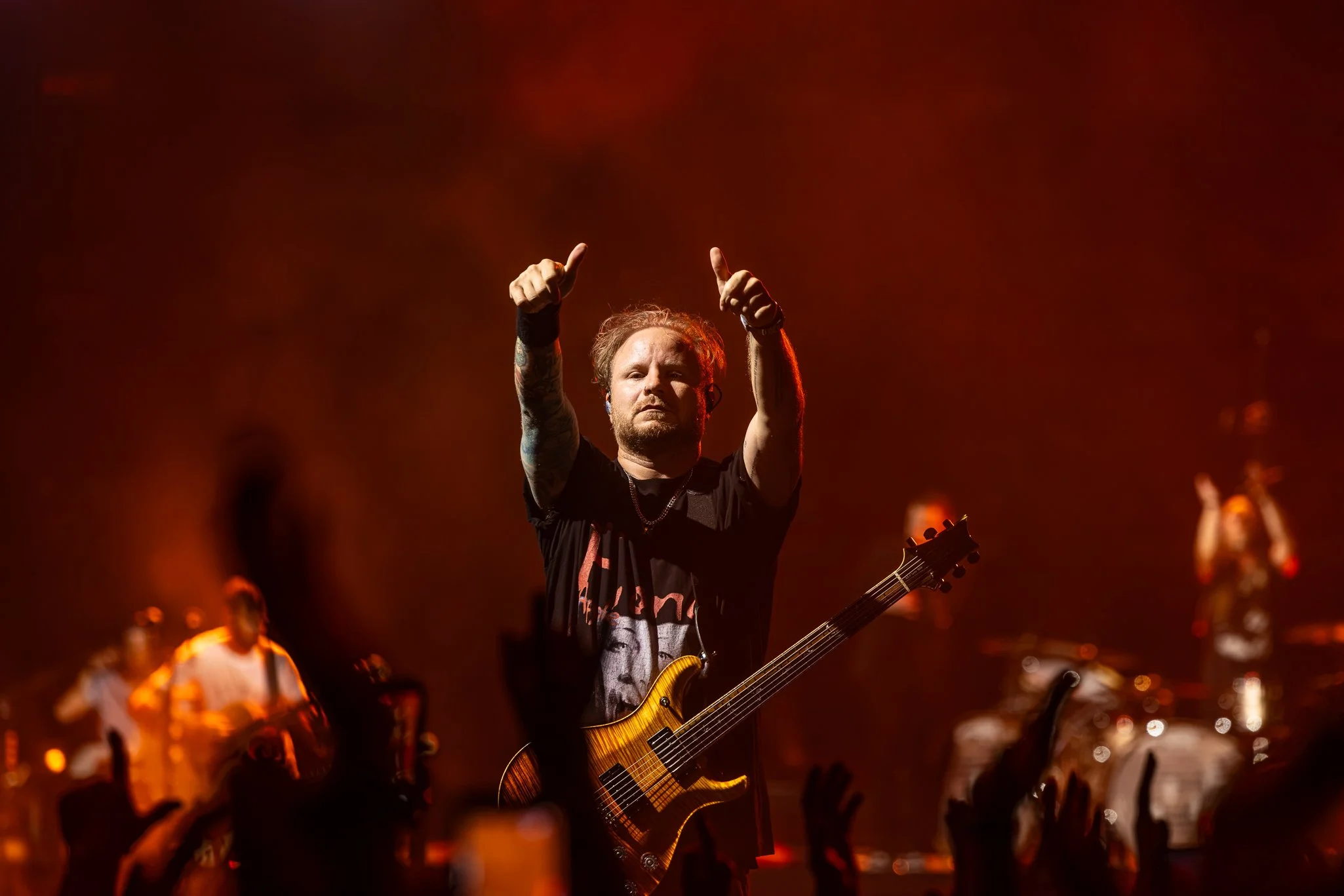 Male musician on stage playing an electric guitar with a dark background and red lighting, cheering along with audience.