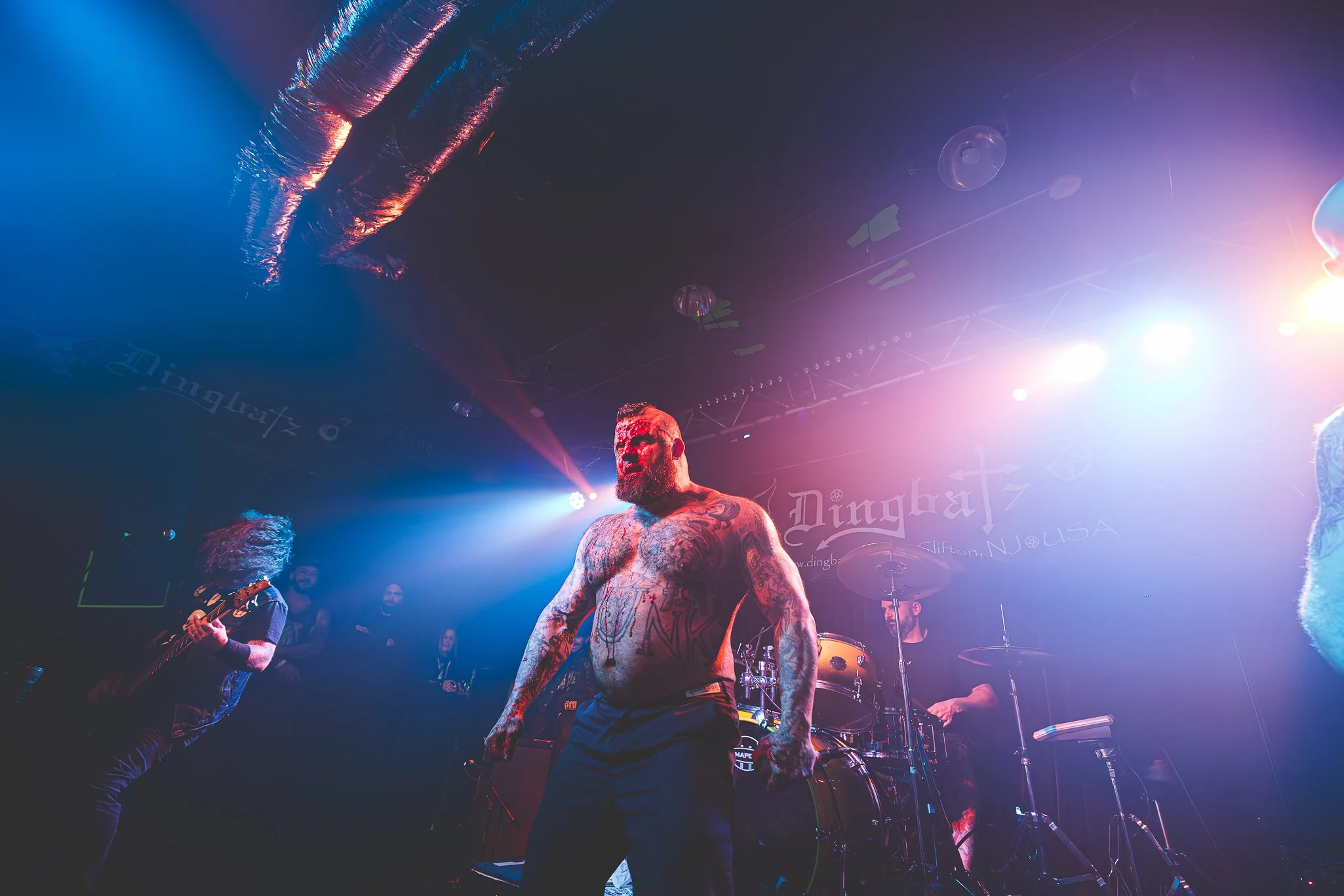 A tattooed shirtless male musician with a beard performing on stage in a concert venue with colorful lighting.