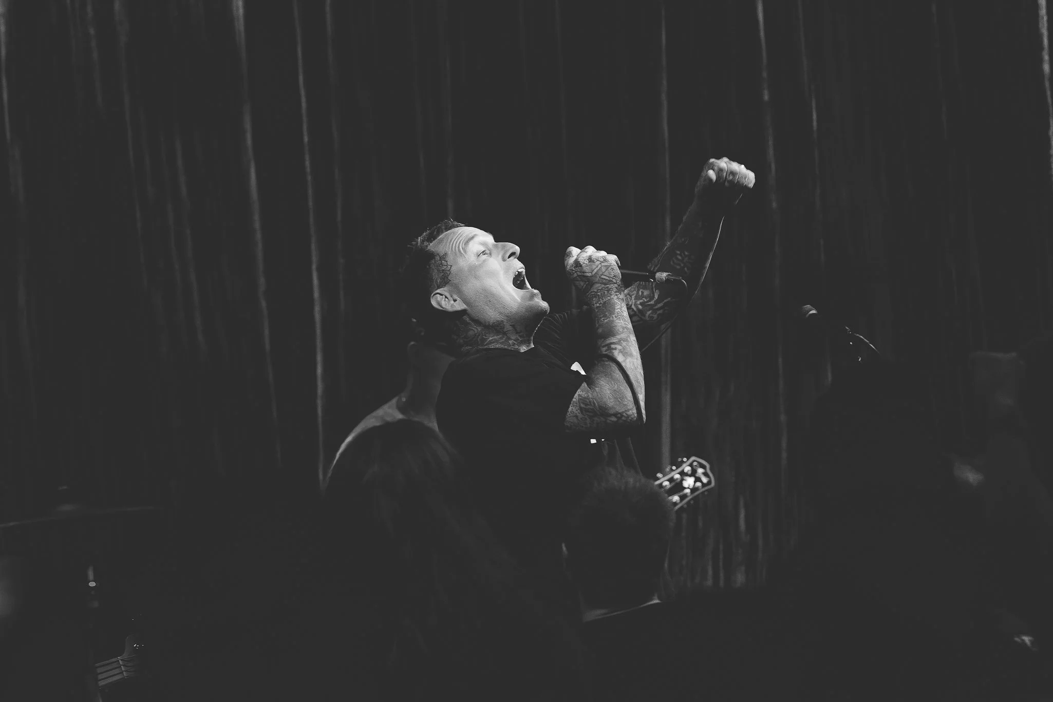 A person with tattoos singing into a microphone while playing an acoustic guitar during a rainstorm at night.