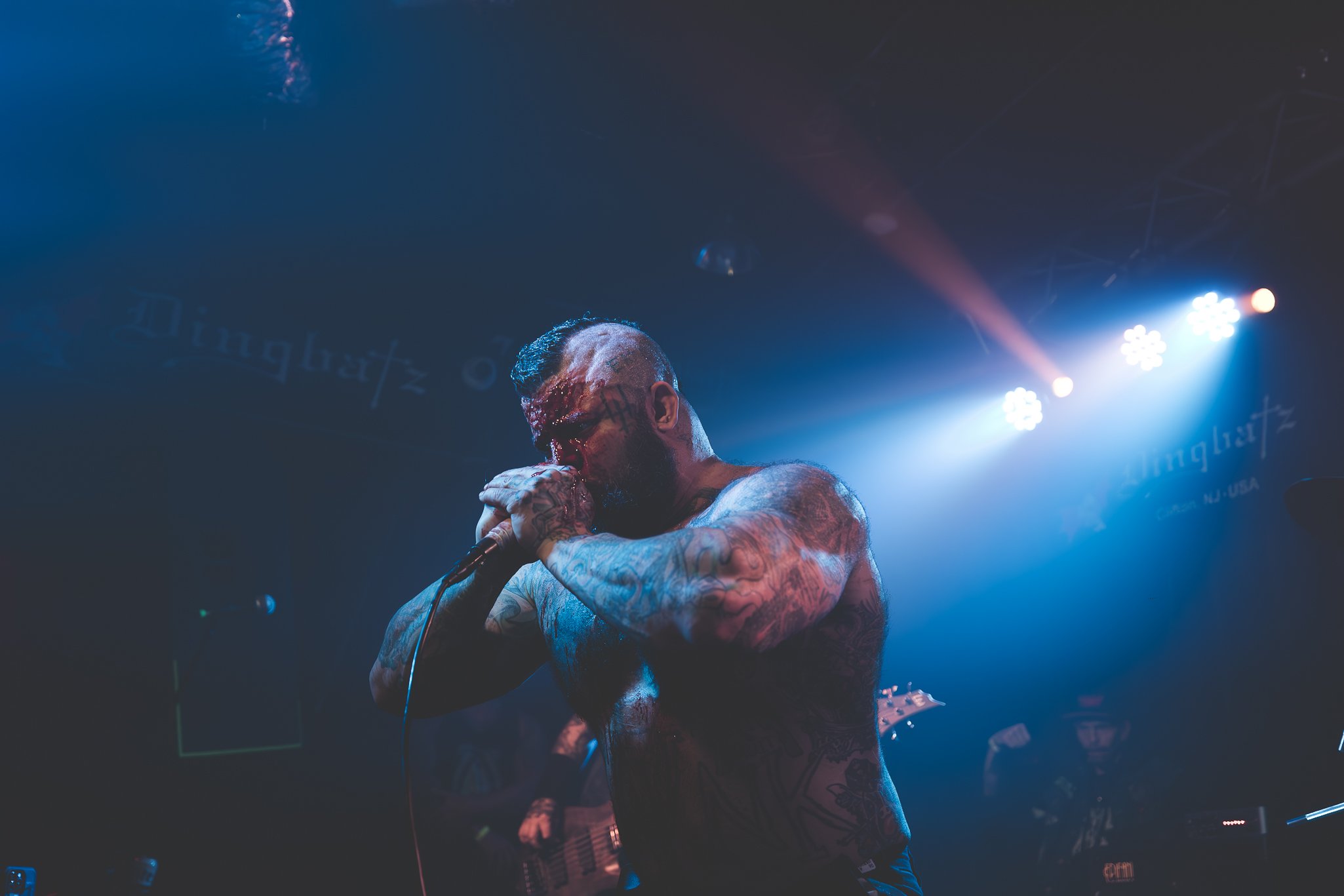 A tattooed male singer performs on stage, holding a microphone with both hands near his mouth, with stage lights illuminating him and a dark background featuring faint text.