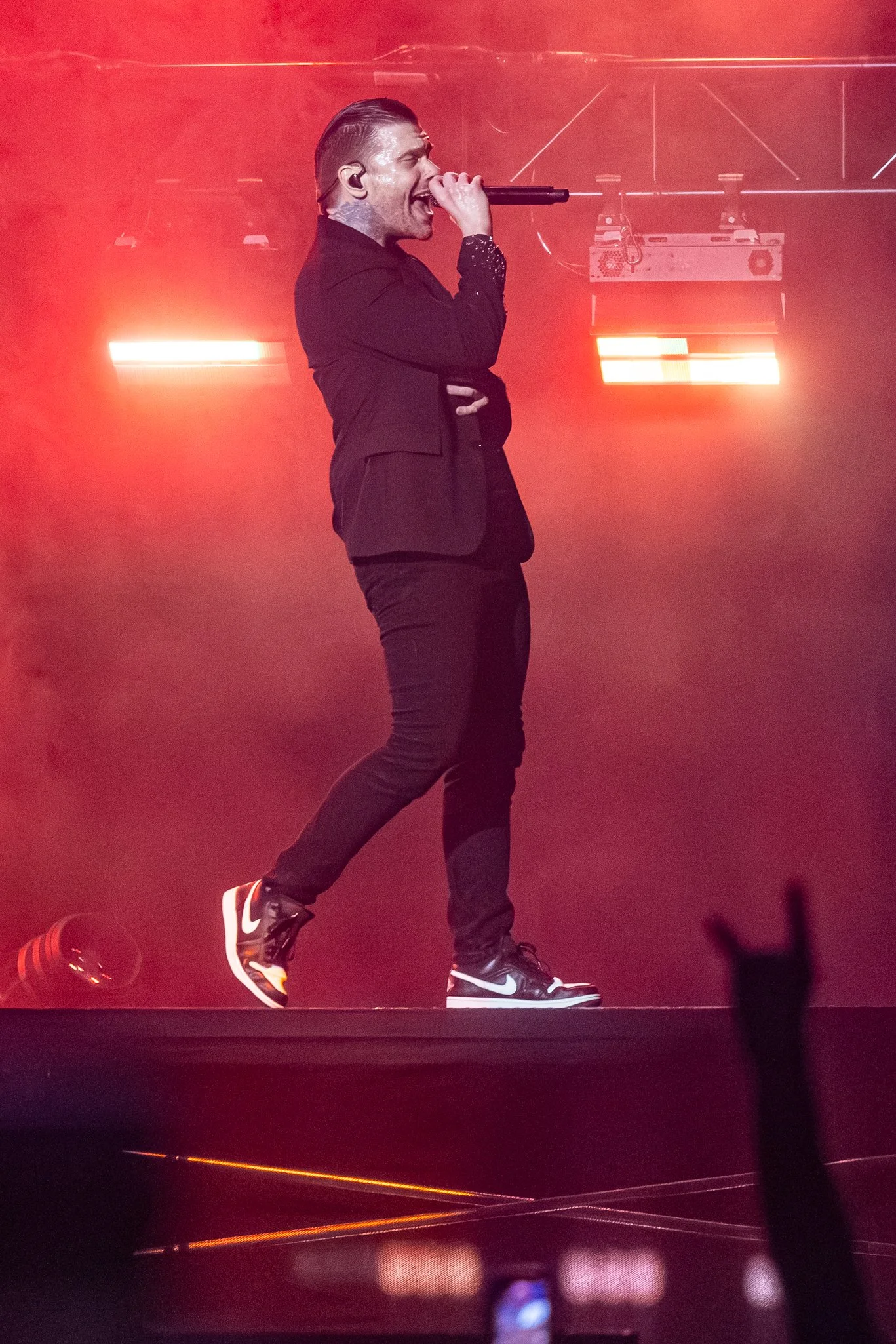 Male singer performing on stage with a microphone, wearing black clothing and sneakers, with stage lighting and an audience in the background.