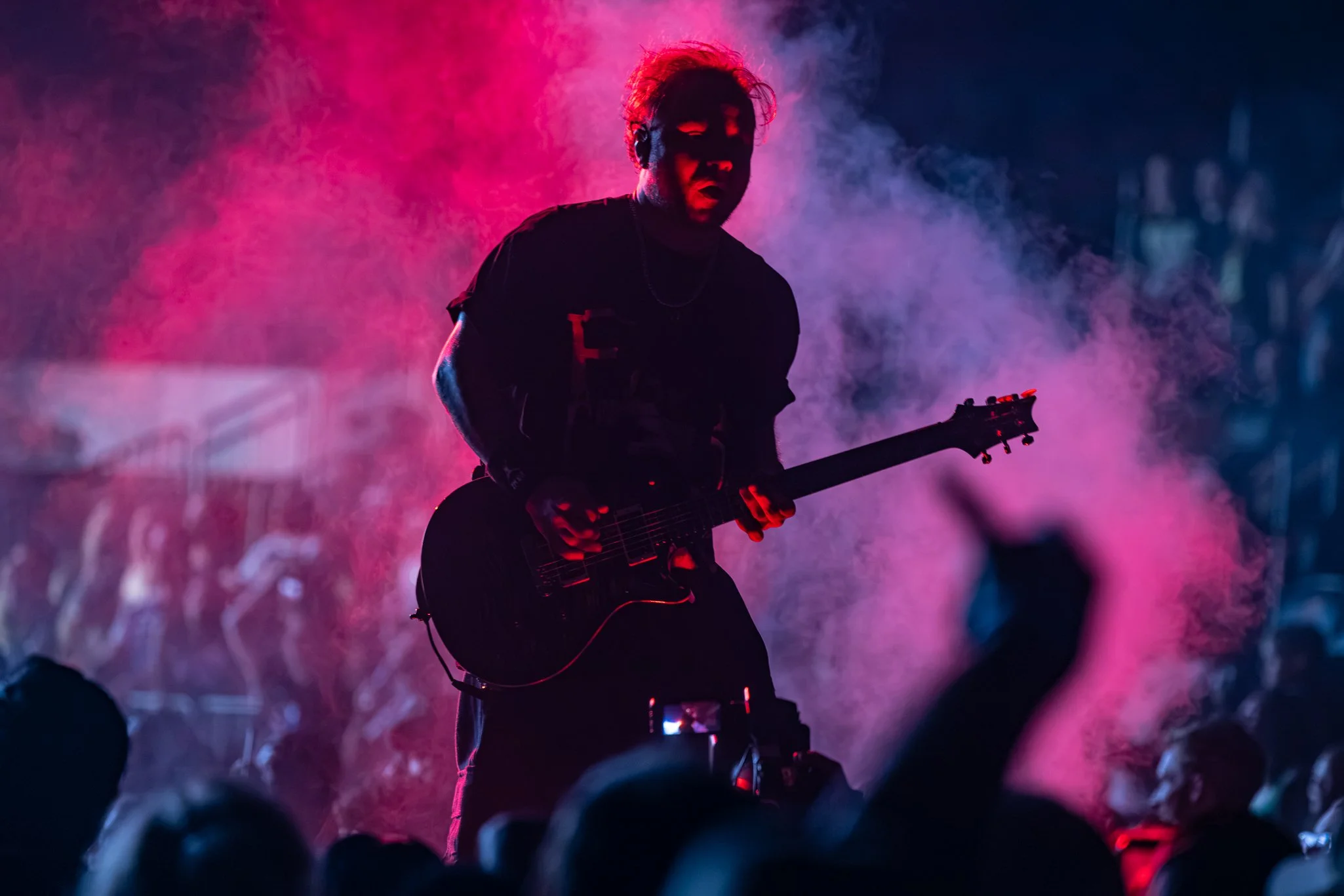 A musician playing an electric guitar on stage surrounded by colorful pink and blue smoke, with a dark background and audience in the foreground.