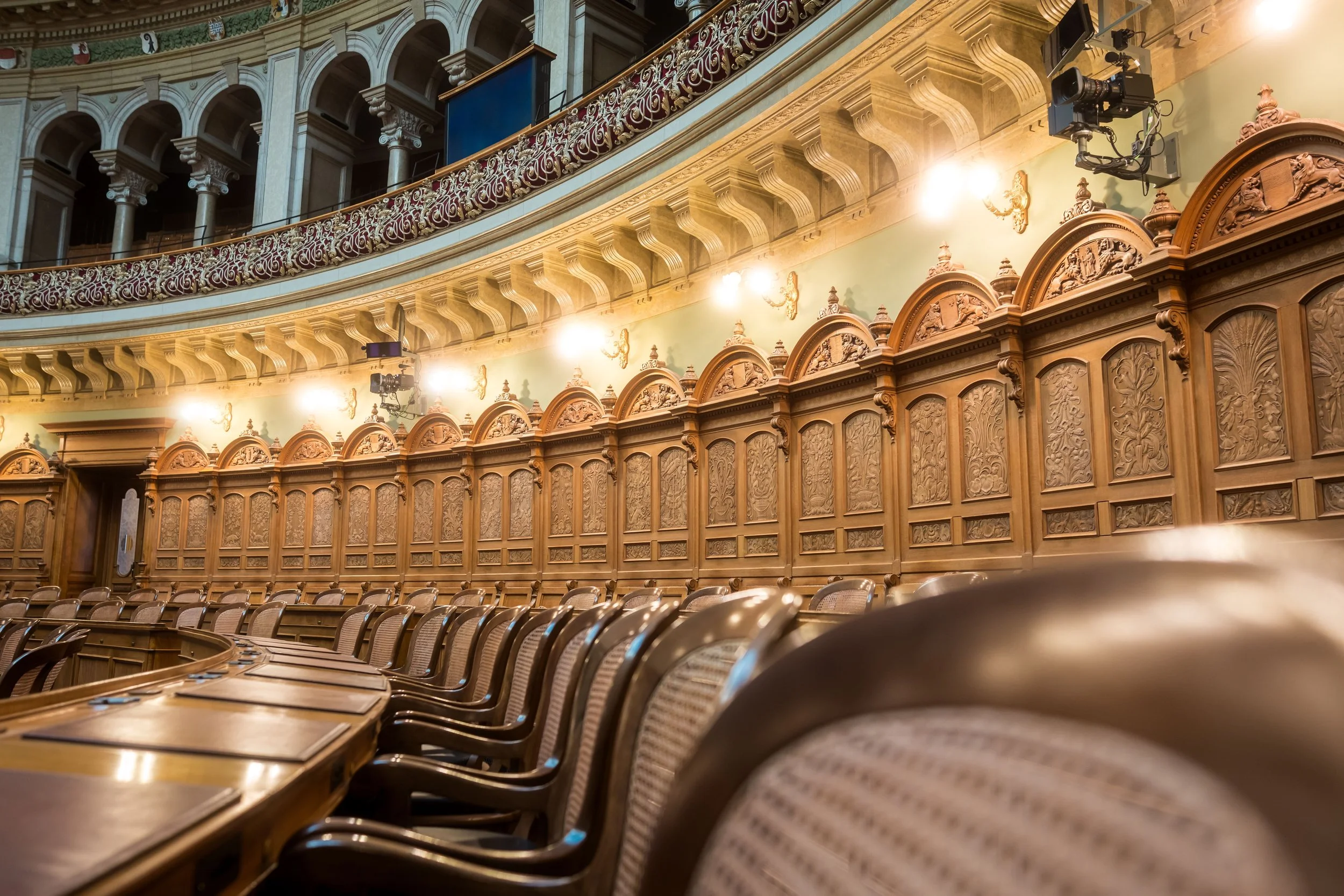 View of an empty ornate theater or auditorium with wooden paneling, decorative moldings, leather seats, and chandeliers.